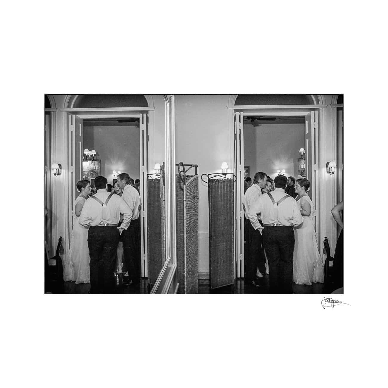 Black and white image of a bride and groom talking with groomsmen in a hallway. They are reflected in a mirror on the right side. The setting appears to be a classic or vintage style venue.