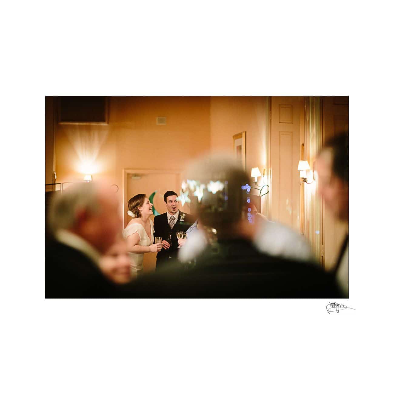 A bride and groom stand together, smiling and holding glasses, in a warmly lit room. Blurred figures appear in the foreground, adding depth to the wedding scene.