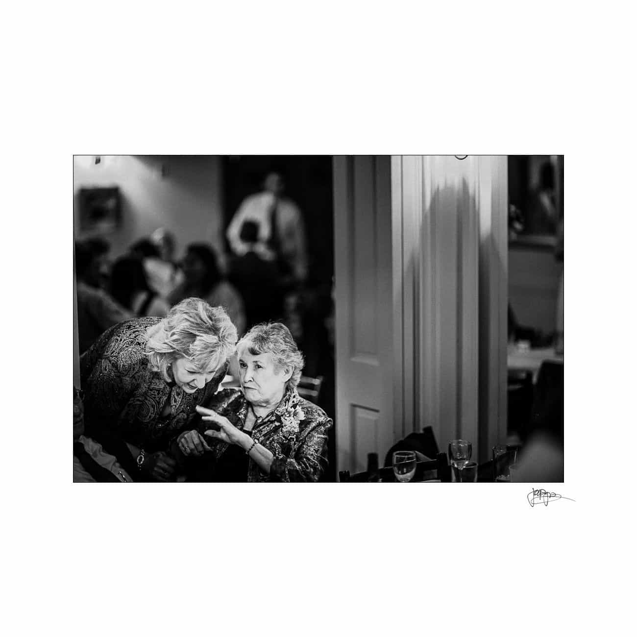 A black and white photo of two women, one elderly with short hair and the other with medium-length hair, engaged in conversation at a crowded indoor gathering. The elderly woman gestures with her hand, while the other leans in to listen.