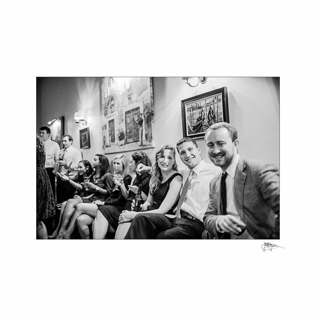 A black and white photo of a group of people seated in a warmly lit room, socializing and smiling. The setting appears to be a casual gathering or event. Artwork is displayed on the wall behind them.