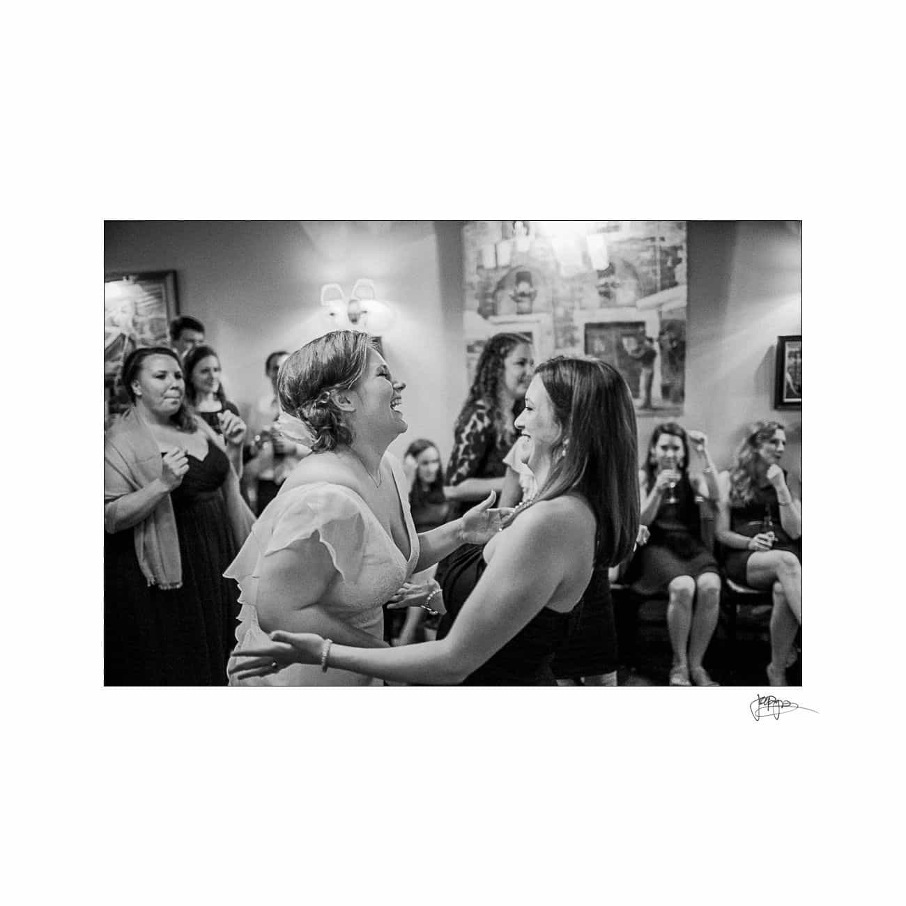 A black and white image of two women joyfully dancing together in a room. They are surrounded by a crowd of people watching and smiling. Decorations and portraits adorn the walls, creating a lively atmosphere.