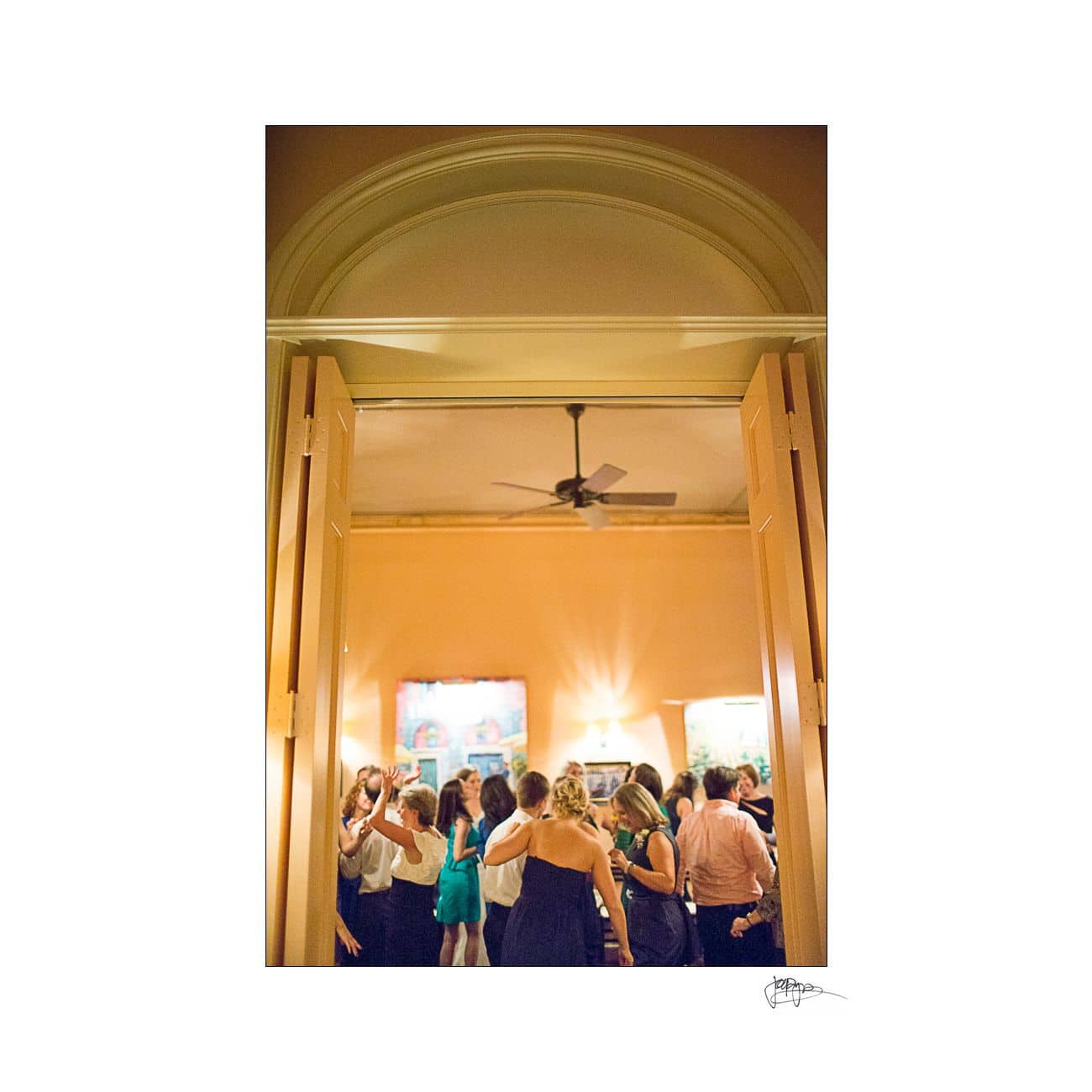 A group of people socializing in a warmly lit room with high ceilings. The room has large open double doors and artworks on the walls. A ceiling fan is visible above the gathering.