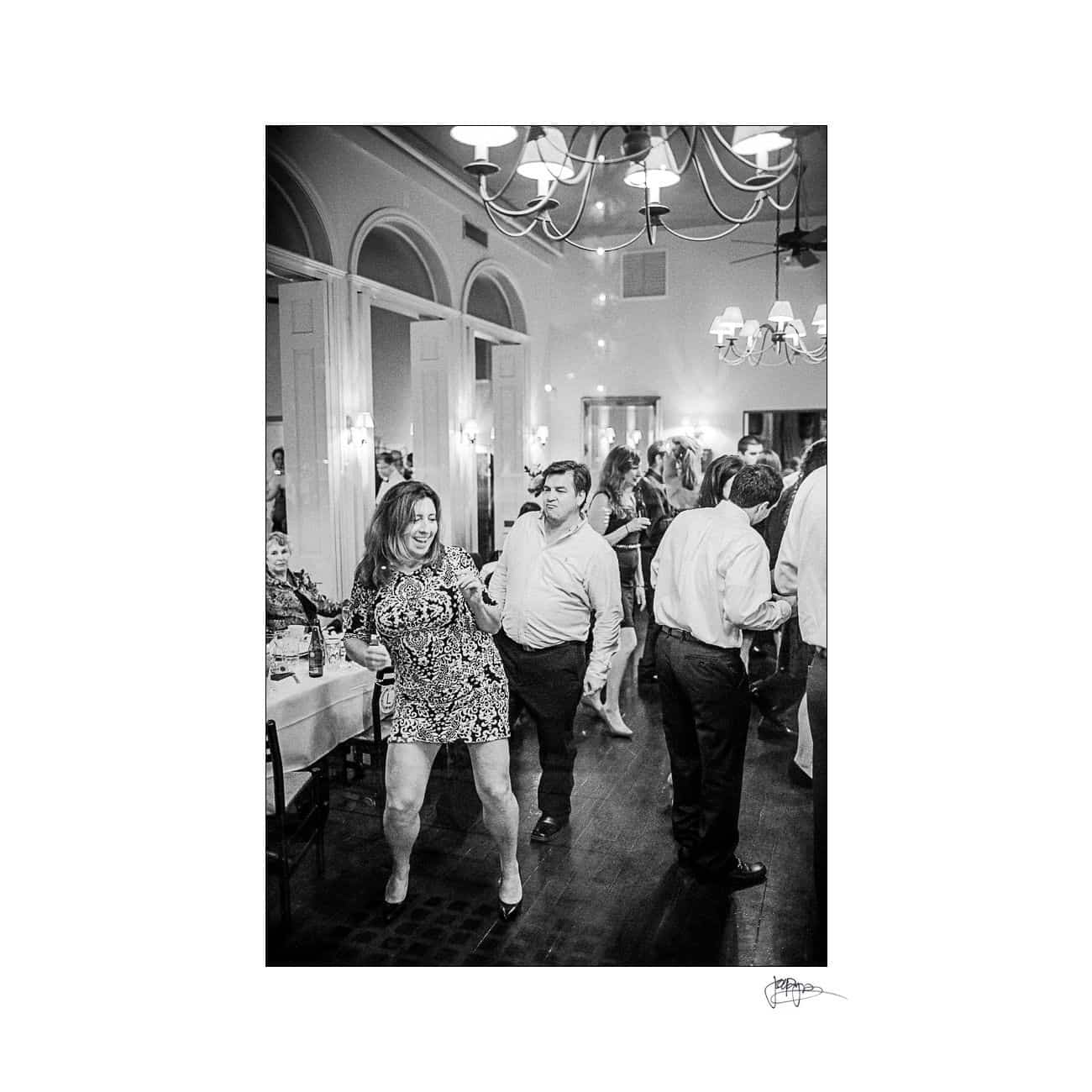 A black and white photo of people dancing at a party. A woman in a patterned dress dances energetically in the foreground, while men in shirts and trousers dance in the background. Tables and chandeliers are visible above.