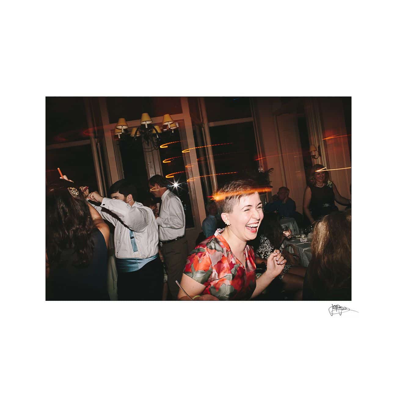 A person in a colorful dress is laughing joyfully at an indoor party. Other guests are dancing and mingling in semi-formal attire. Strings of light add a lively atmosphere to the scene.