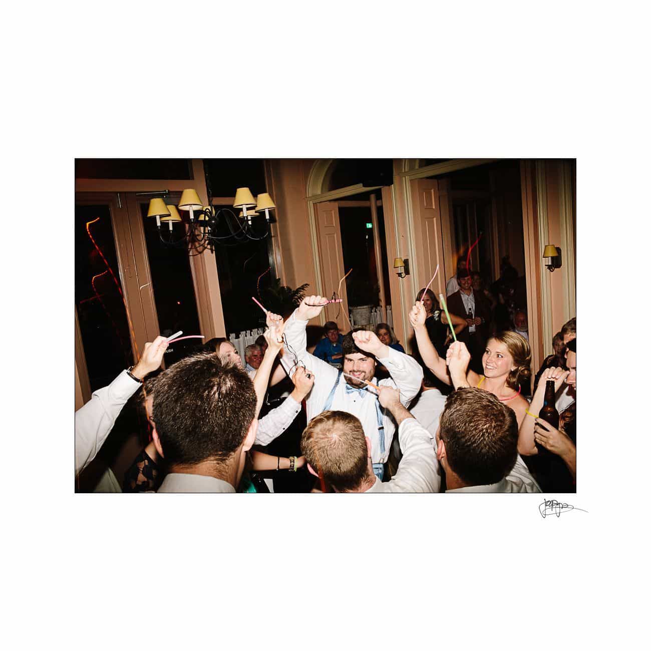 A group of people raising sparklers in a celebratory manner indoors. A man in the center, wearing a shirt and tie, is smiling. The room has large windows and warm lighting, creating a festive atmosphere.