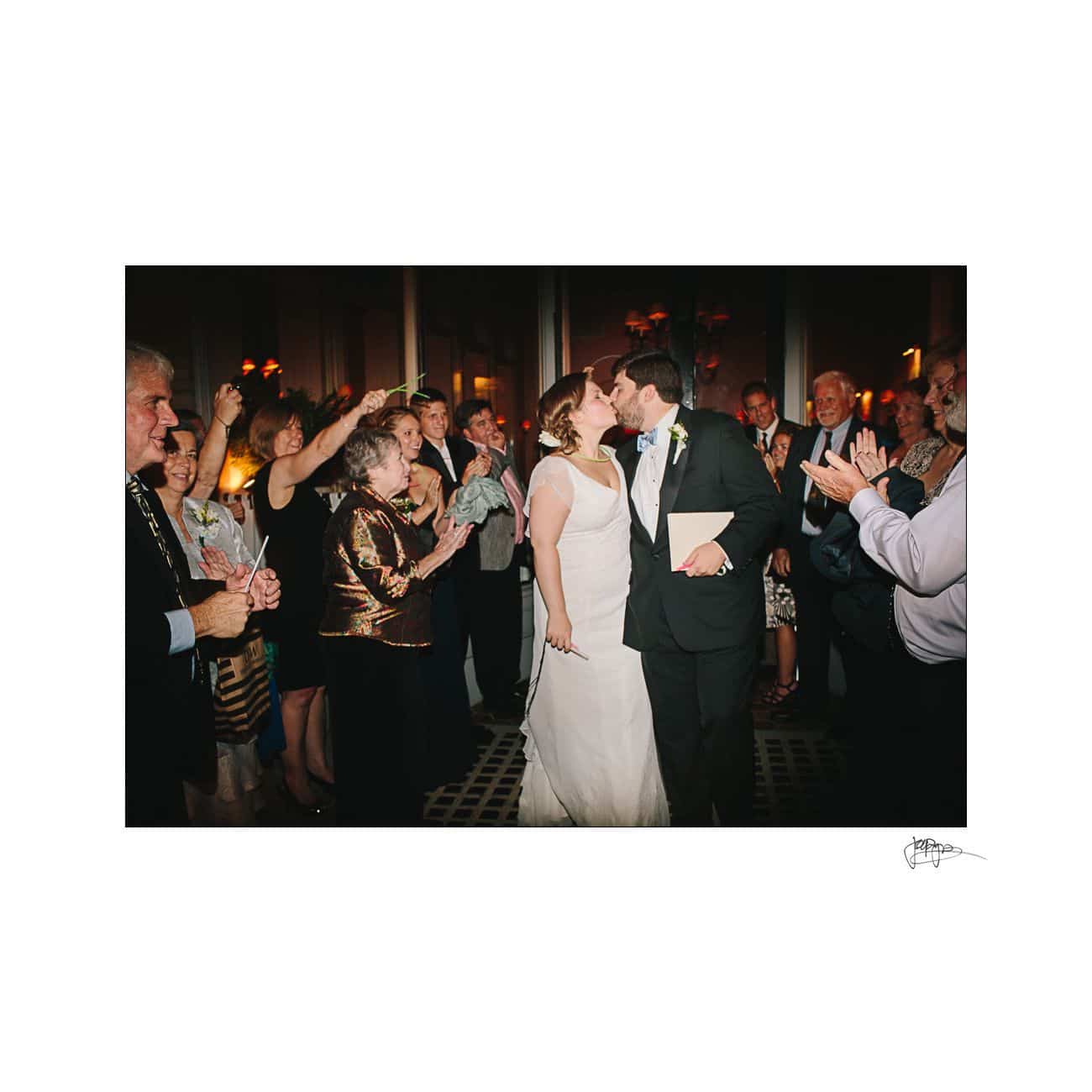 A bride and groom share a kiss while surrounded by a cheering crowd at their wedding reception. The bride wears a white gown, and the groom is in a suit. Guests are clapping and celebrating the couples special moment.