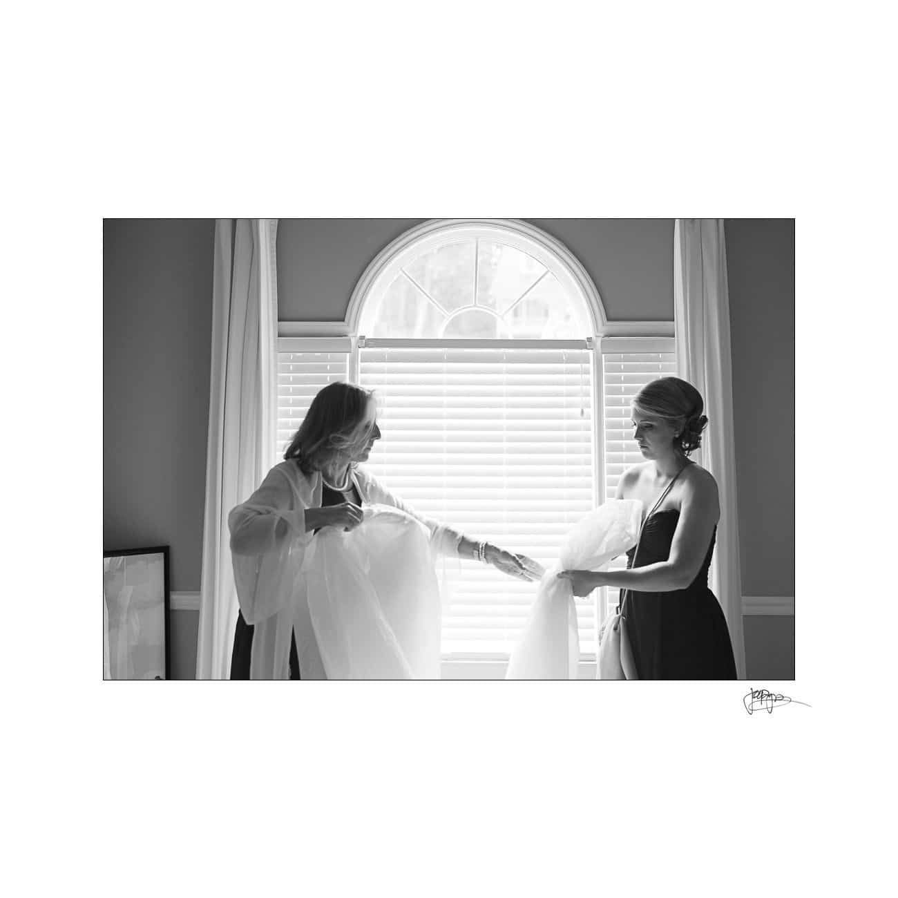 A black and white photo of two women standing by a window with closed blinds. They appear to be arranging a piece of tulle or fabric together. Sunlight filters through, creating a soft glow around them.