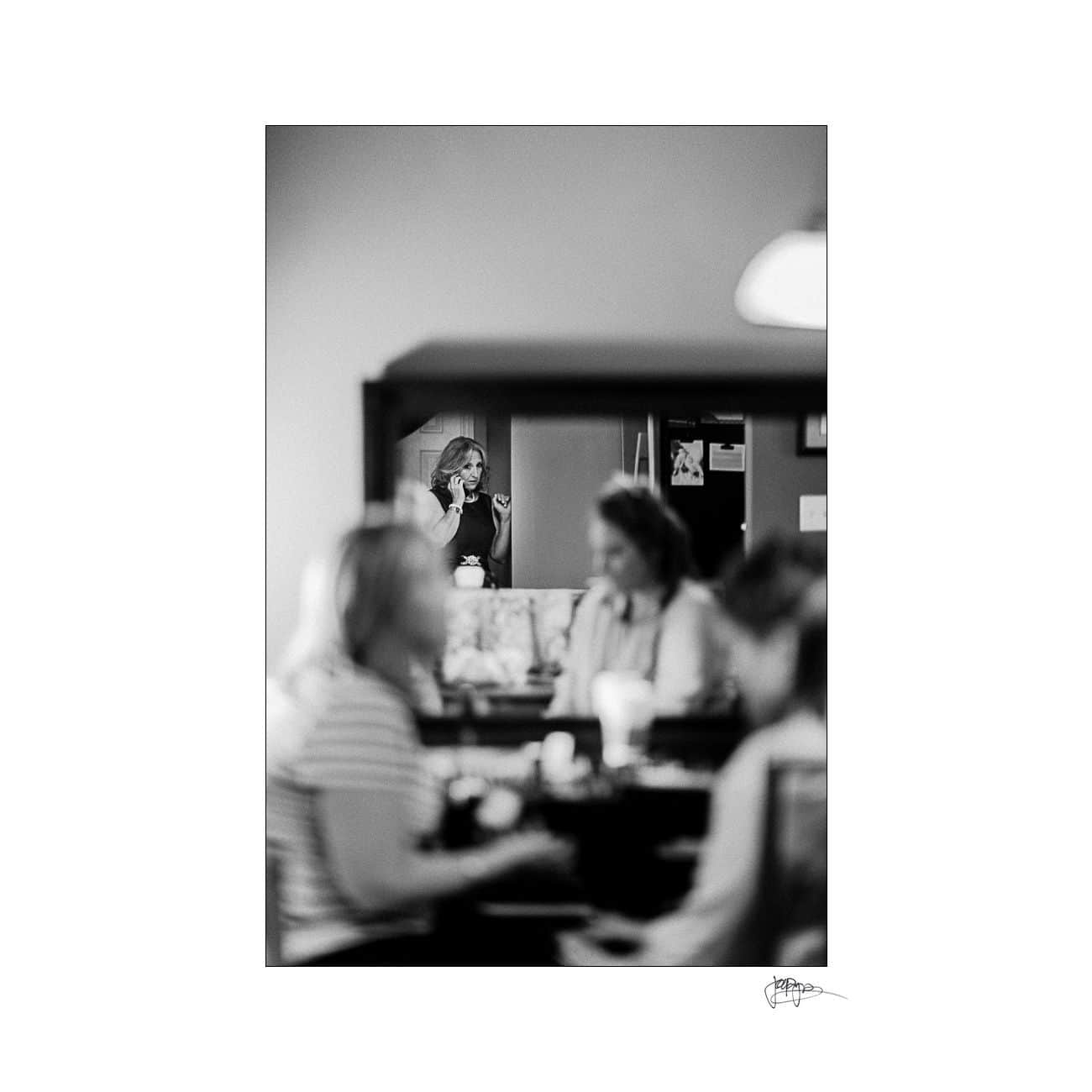 A black and white photo depicts a reflection of a woman taking a picture in a mirror. In the foreground, two blurred figures are sitting at a table, engaged in a conversation.