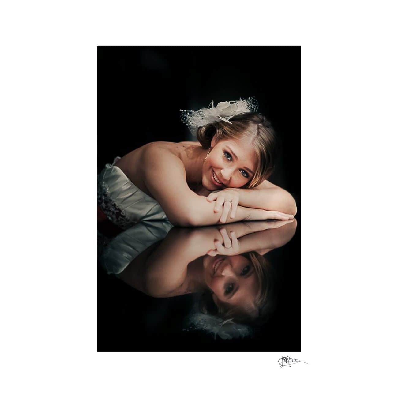 A woman in a white dress lies on a reflective surface, her chin resting on her folded hands. She has a feathered hair accessory and smiles at the camera, with her reflection visible below. The background is black.