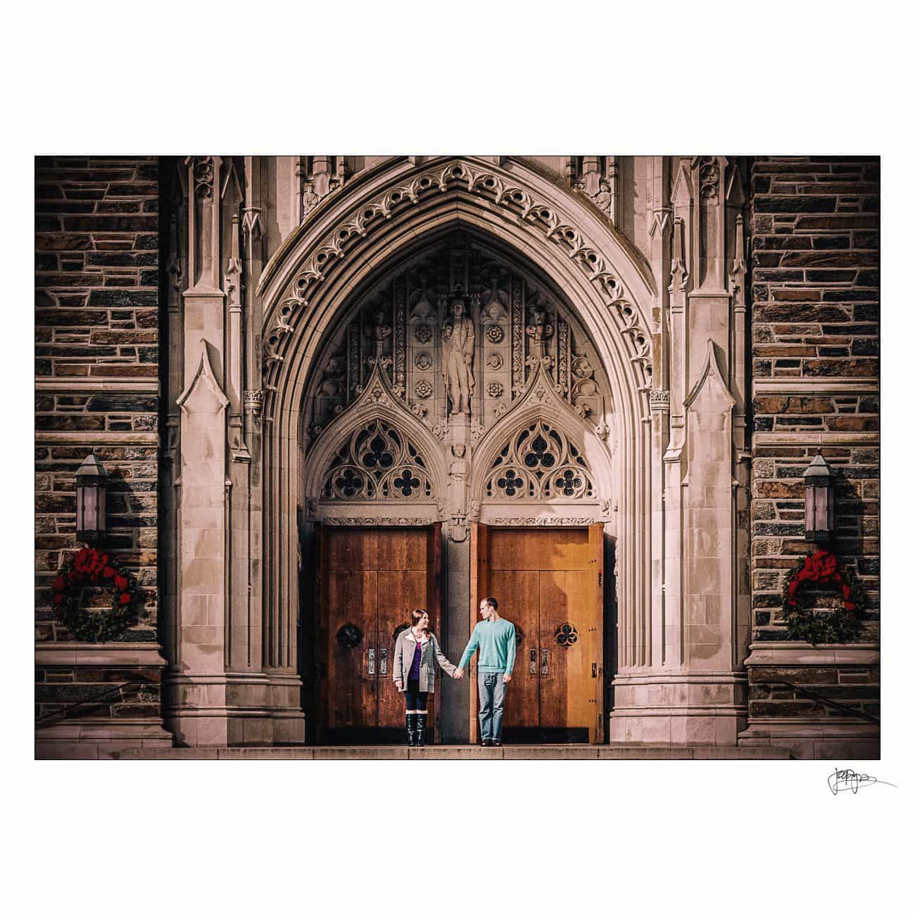 Two people stand holding hands in front of large wooden double doors with ornate stone arches, set in a gothic-style building at Duke University—an elegant setting perfect for engagement photography, with lanterns and red wreaths on either side.