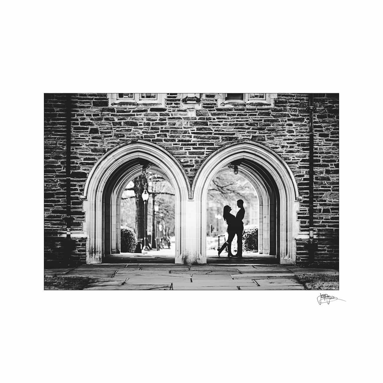 A black and white photo of two people facing each other under adjoining archways in a brick wall. The arches cast shadows on the ground, and trees are visible in the background through the open spaces.
