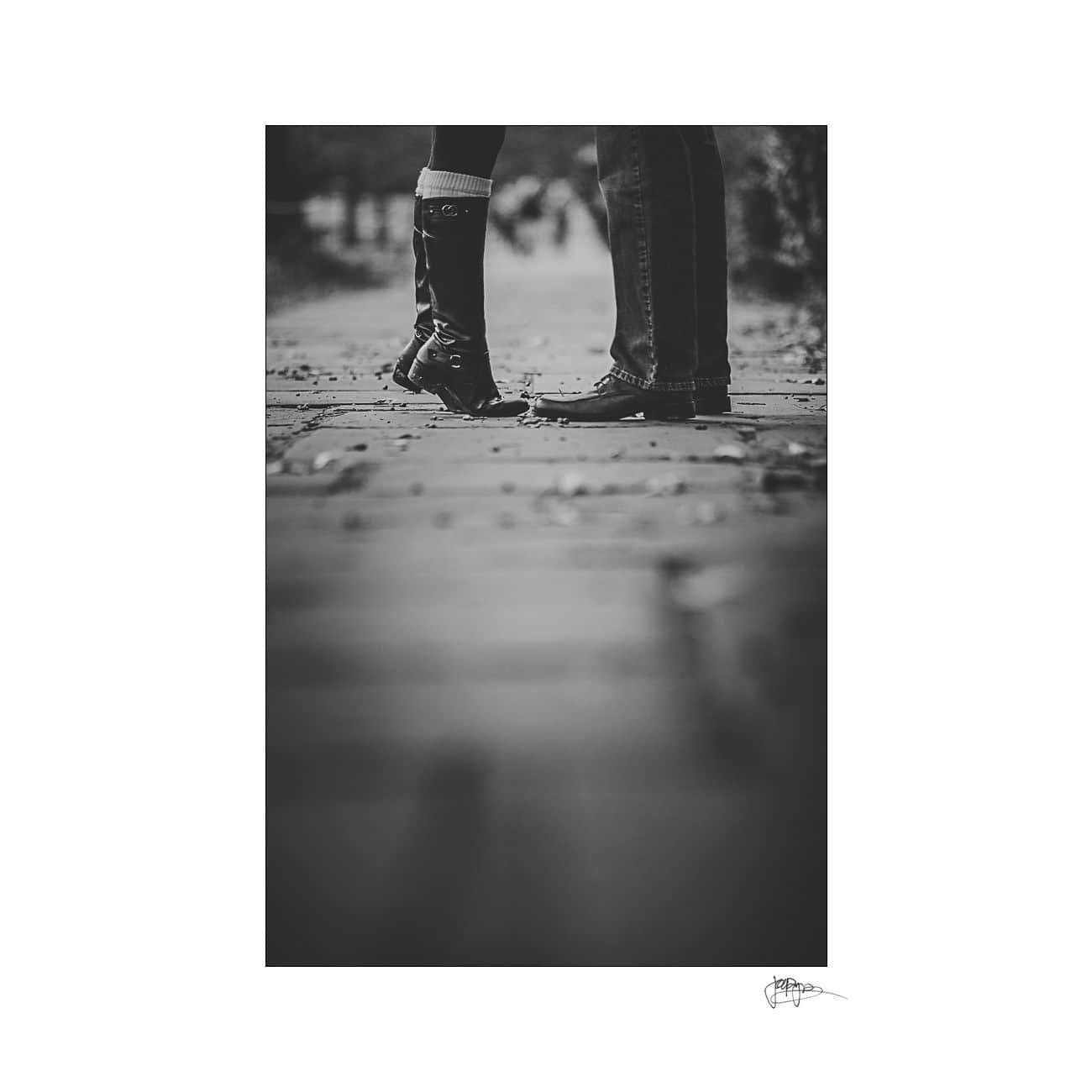 Black and white photo of two people from the knees down, standing on a paved path. One person on tiptoes wearing boots, the other in sneakers. Leaves scattered on the ground, adding a touch of autumn atmosphere.