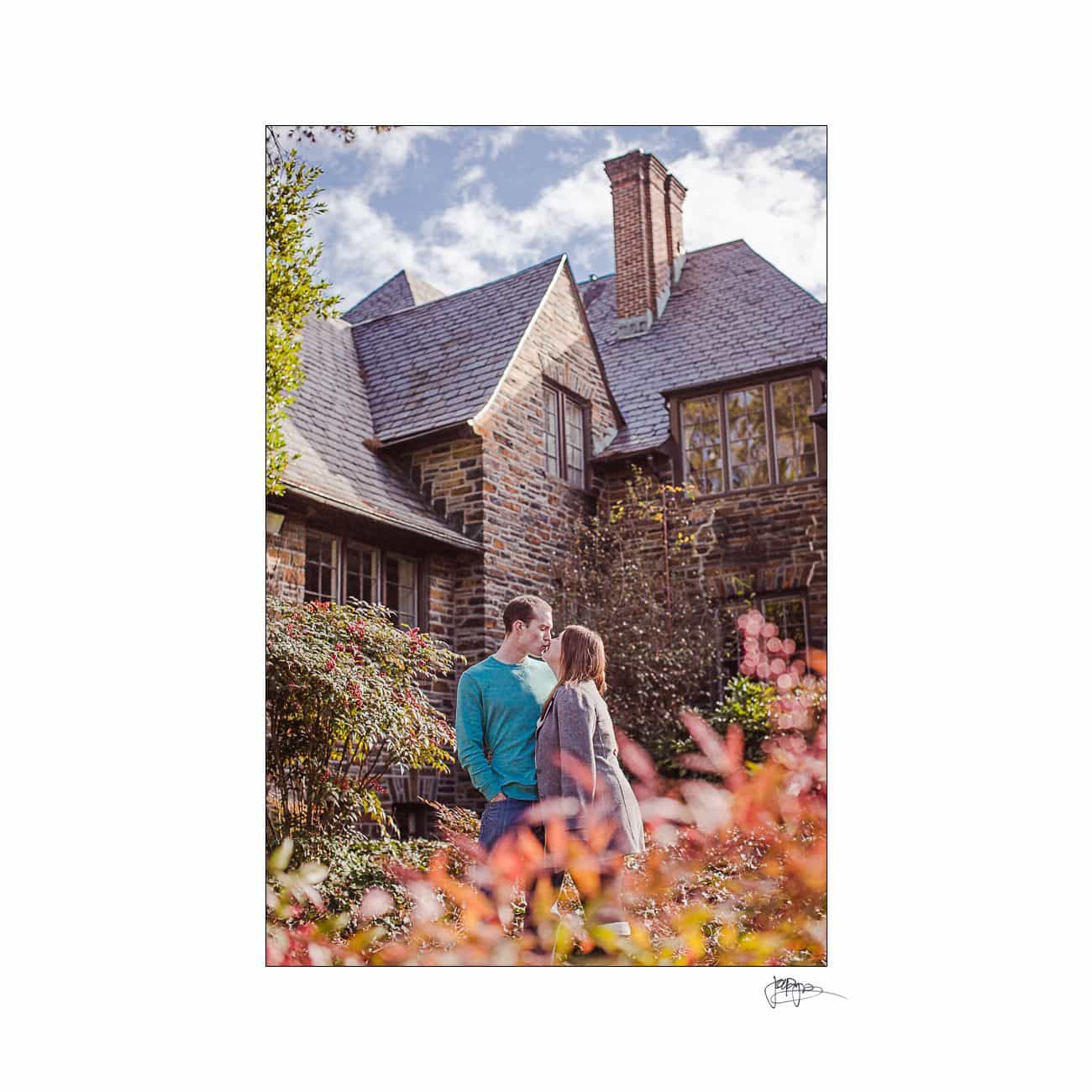A couple stands in front of a quaint, stone house with a steep roof, surrounded by colorful foliage. The sky is partly cloudy, adding a serene backdrop to the autumn scene.