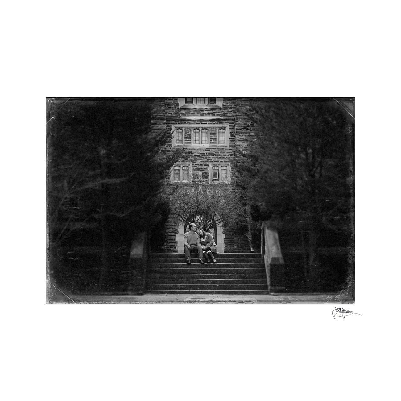 A black and white photo of two people sitting and relaxing on steps in front of a large building. Tall trees line both sides of the stairs, framing the entrance in the background.