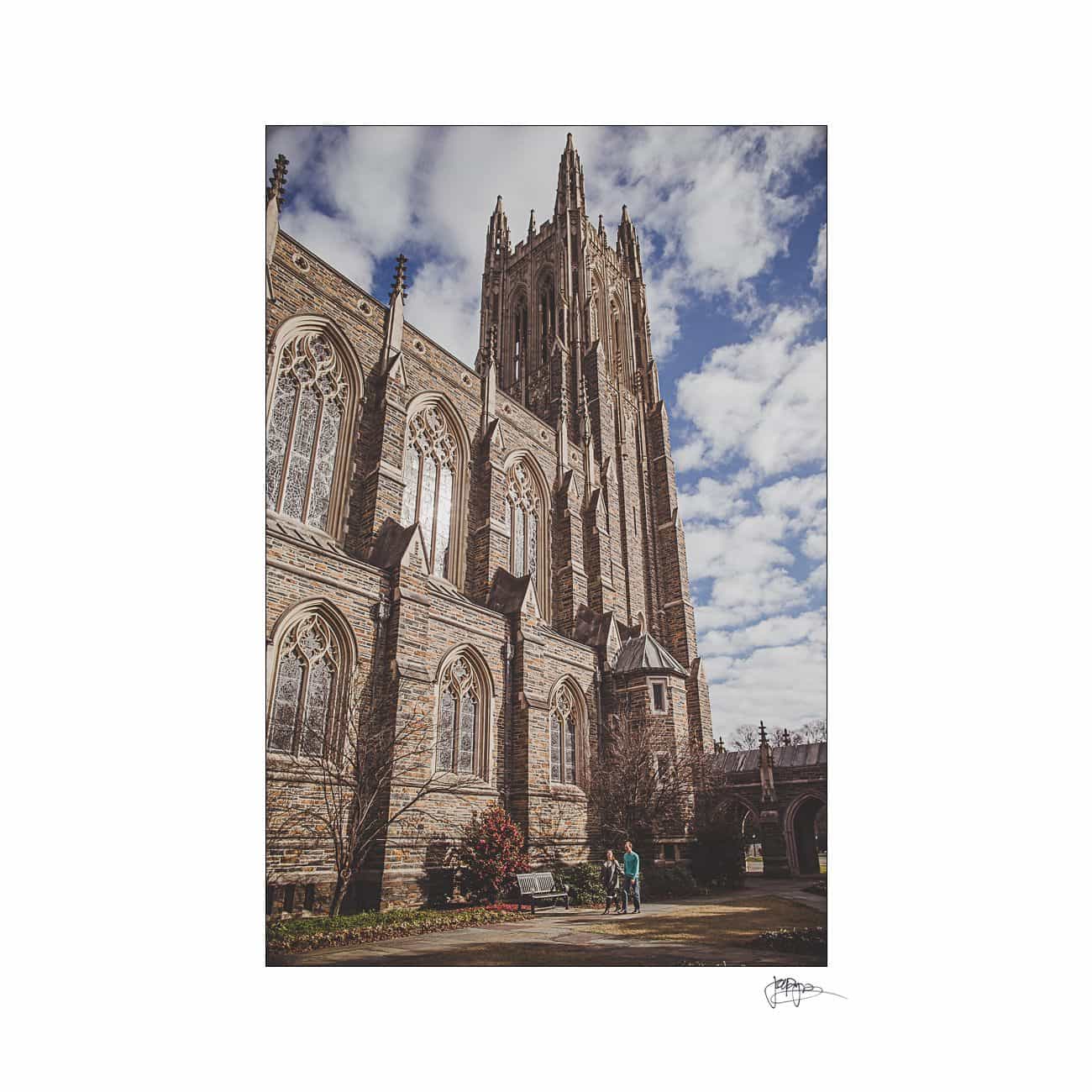 A towering Gothic-style cathedral with intricate stonework stands under a partly cloudy sky. Two people walk along the pathway near the building, surrounded by the lush greenery of the garden.