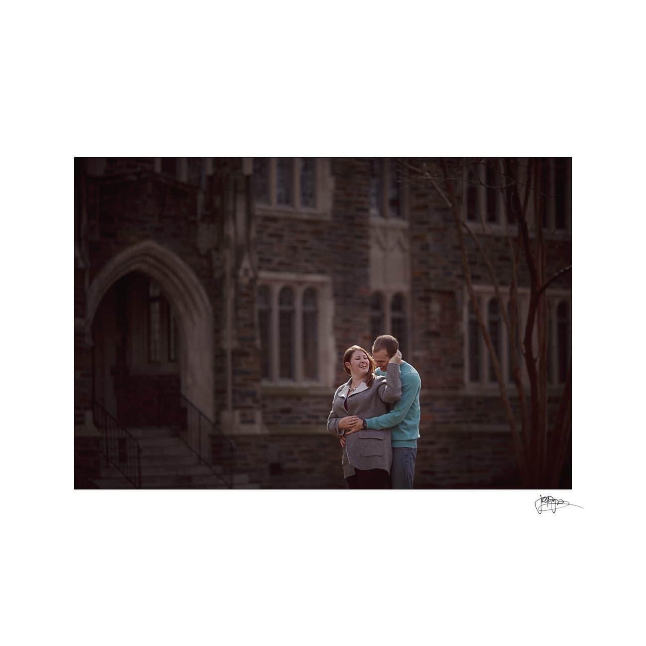A couple stands in an embrace, smiling warmly at each other. The background features a historic building with arched windows and stone walls, adding an architectural charm to the scene.