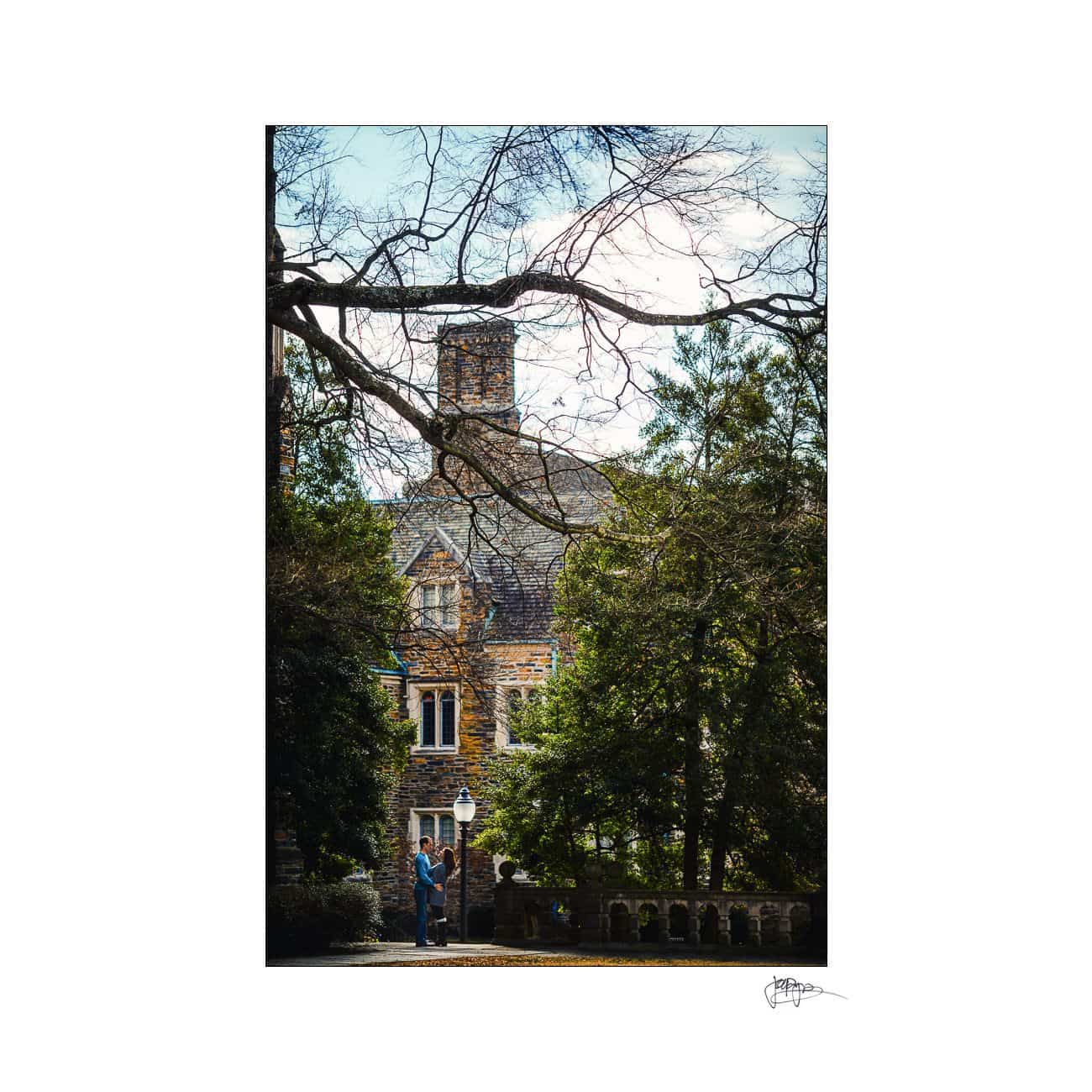 A person stands near a historic stone building partially obscured by trees. The scene is framed by bare branches and evergreen foliage, under a clear sky. A lamp post is visible in the foreground.