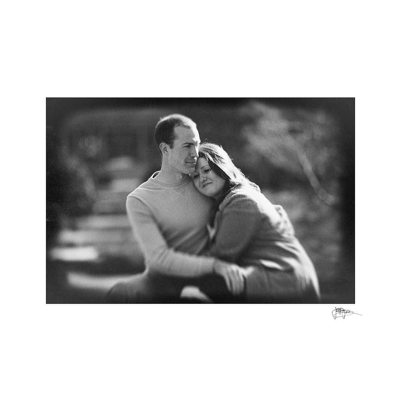 Black and white image of a couple sitting outside. The man is wearing a light sweater and the woman is in a coat, resting her head on his shoulder. They appear content and close, sitting on steps in a blurred outdoor setting.