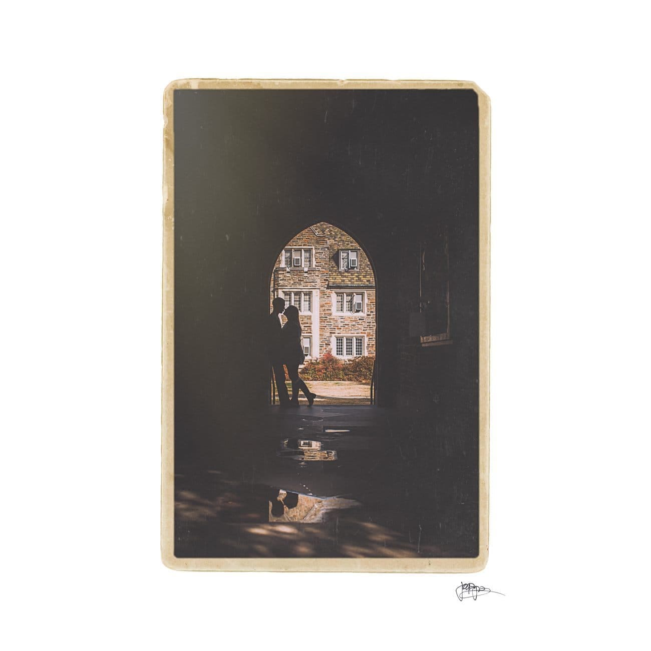 A silhouette of a couple embracing under an arched stone walkway, with a historic building featuring large windows in the background. A small puddle reflects the arch and couple, adding a dreamy effect to the scene.