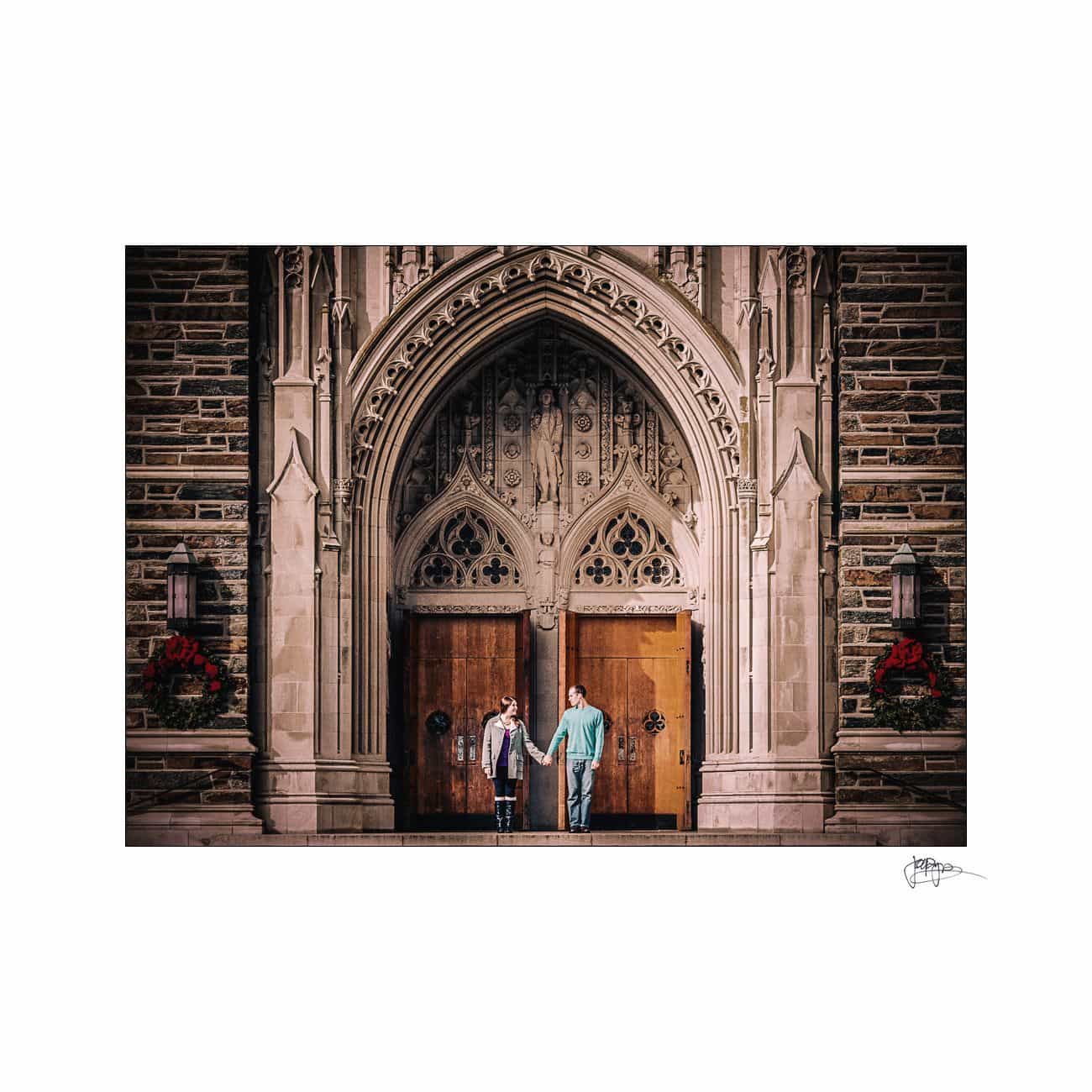 Two people holding hands stand in front of large, ornately carved wooden doors set in a stone building with intricate gothic-style architecture. Red wreaths hang on either side of the doorway.
