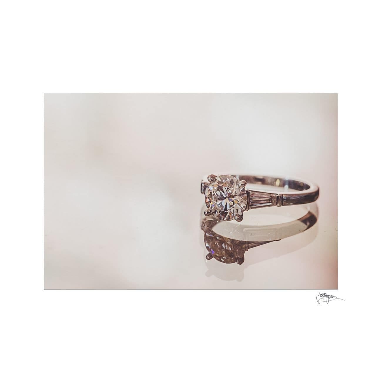 A close-up of a diamond ring with a reflective surface. The band is silver, featuring a large central diamond with smaller stones on the sides. The background is a soft, blurred cream color.