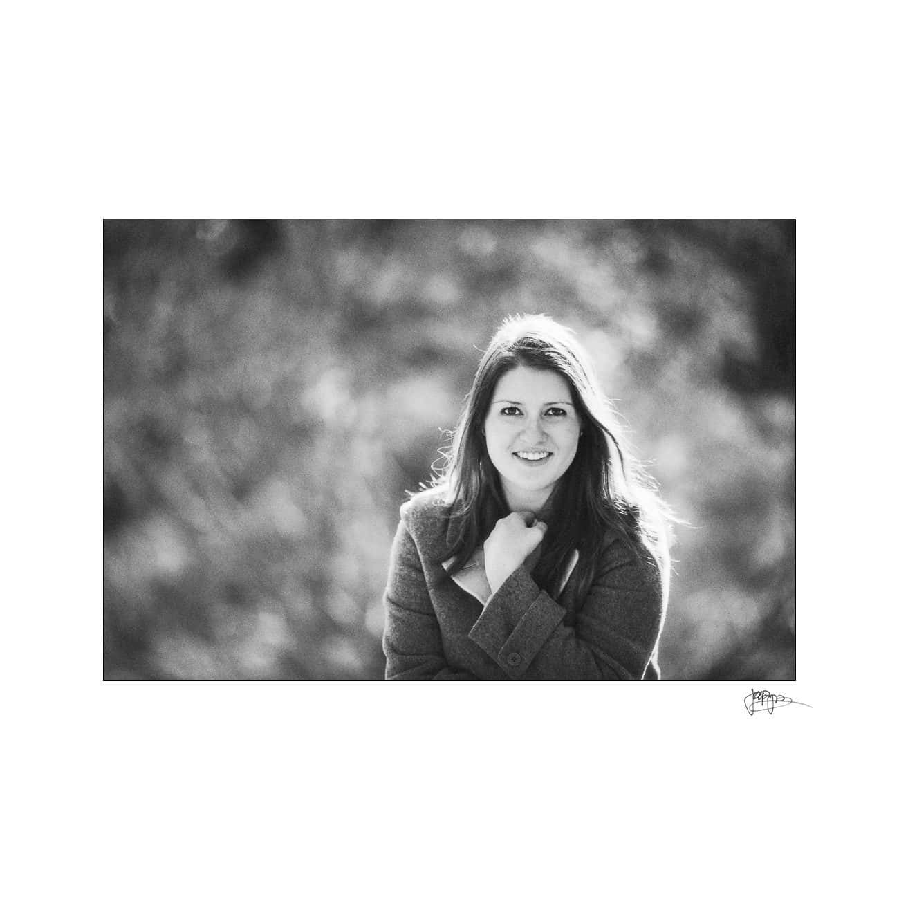 A black and white photo of a woman smiling warmly at the camera. She is standing outdoors, wearing a coat. The background is softly blurred, adding a serene and relaxed ambiance to the image.
