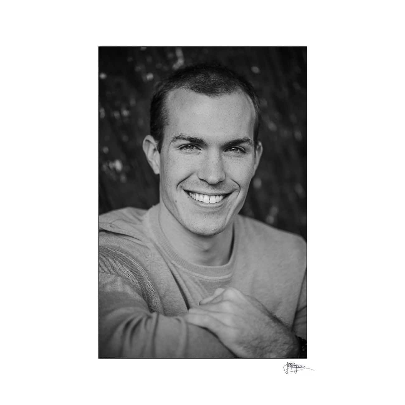 A black and white portrait of a smiling man with short hair. He is wearing a light-colored sweater and has his arms crossed in front of him, looking at the camera. The background is blurred.