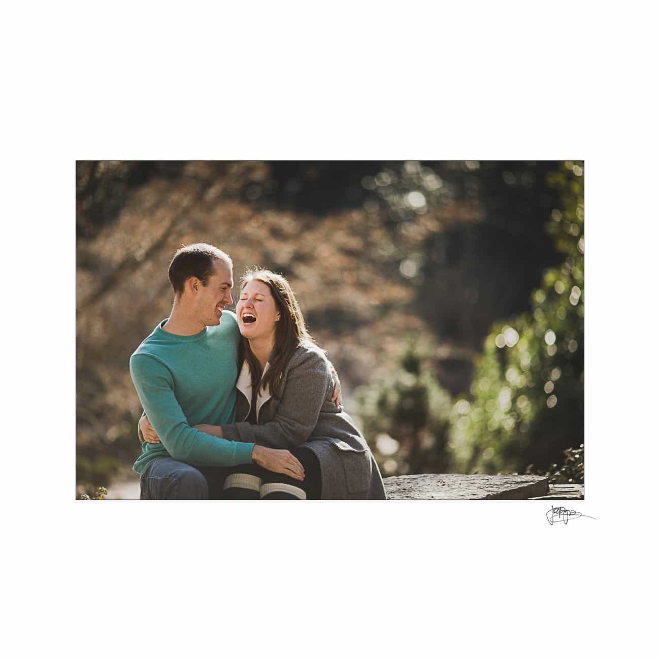 A couple sits outdoors on a sunny day, laughing together. The man wears a turquoise sweater, and the woman has a gray coat. They are embraced, with blurred greenery in the background, creating a warm and joyful atmosphere.