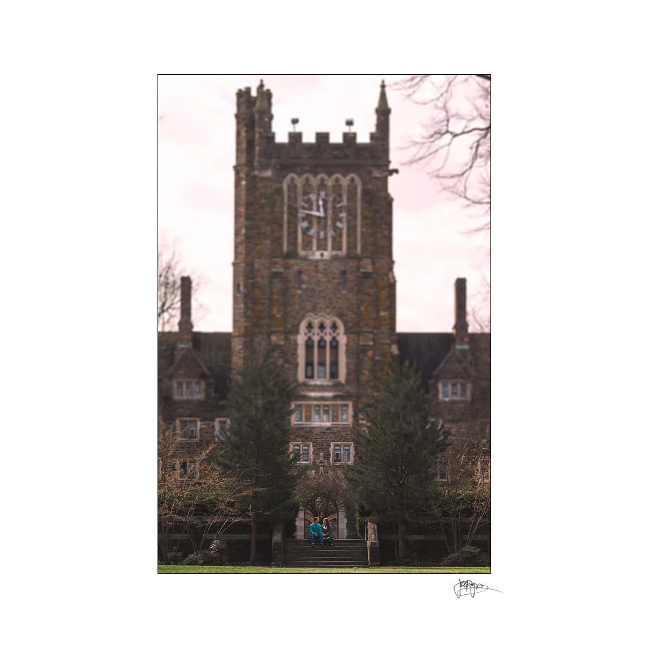 A Gothic-style stone building with a large clock tower and arched windows. Two people stand near the entrance, surrounded by trees and a cloudy sky.