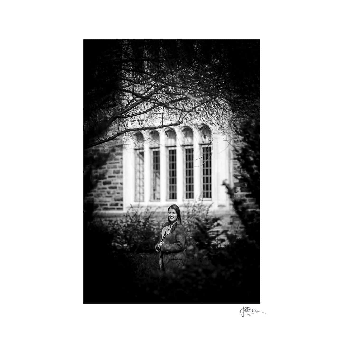 A black and white photograph of a person standing in front of a building with arched windows. They are surrounded by trees and foliage, creating a serene and natural setting. The image has a vignette effect.