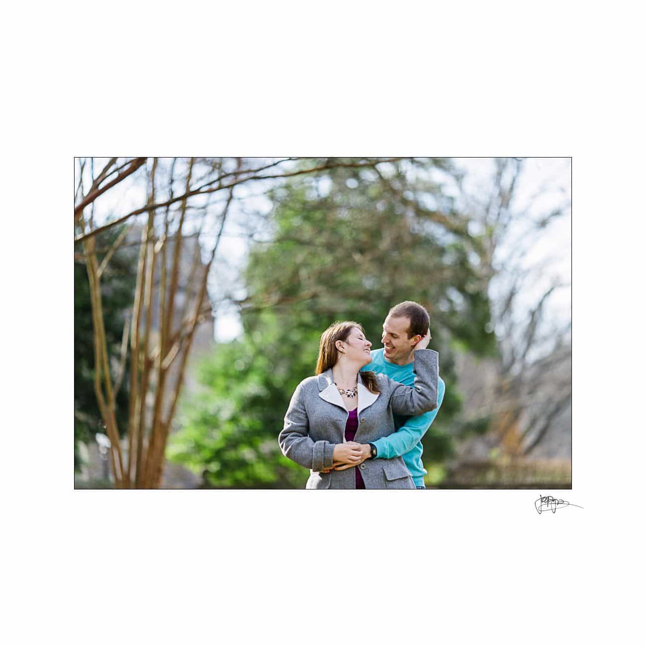 A couple embraces outdoors, smiling at each other. They stand in front of blurred greenery, with bare tree branches visible, suggesting early spring. The woman wears a gray jacket, and the man wears a blue sweater.
