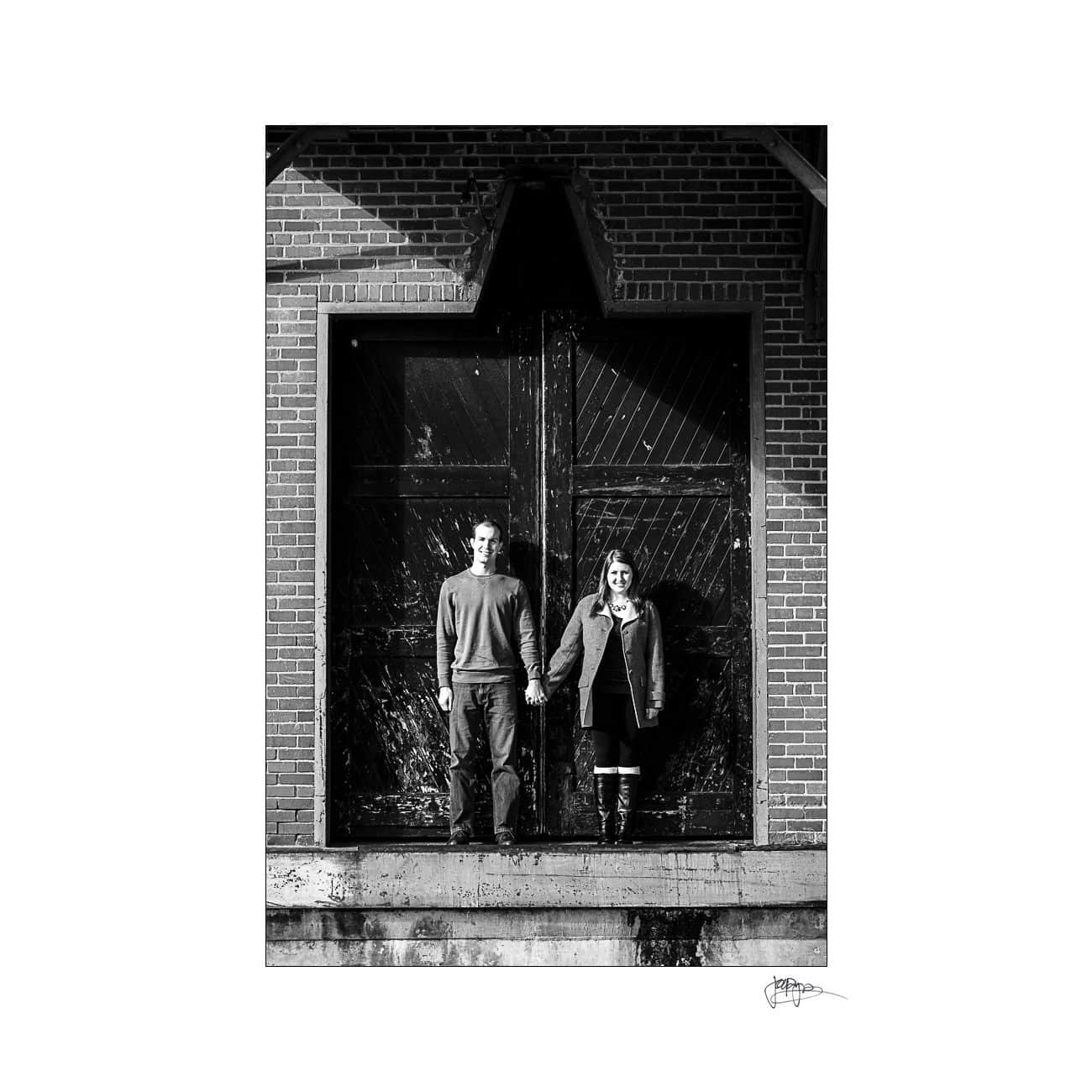 A couple stands holding hands in front of large, weathered wooden doors set in a brick wall. The man on the left and woman on the right are both dressed in casual, dark clothing. The scene is captured in black and white.