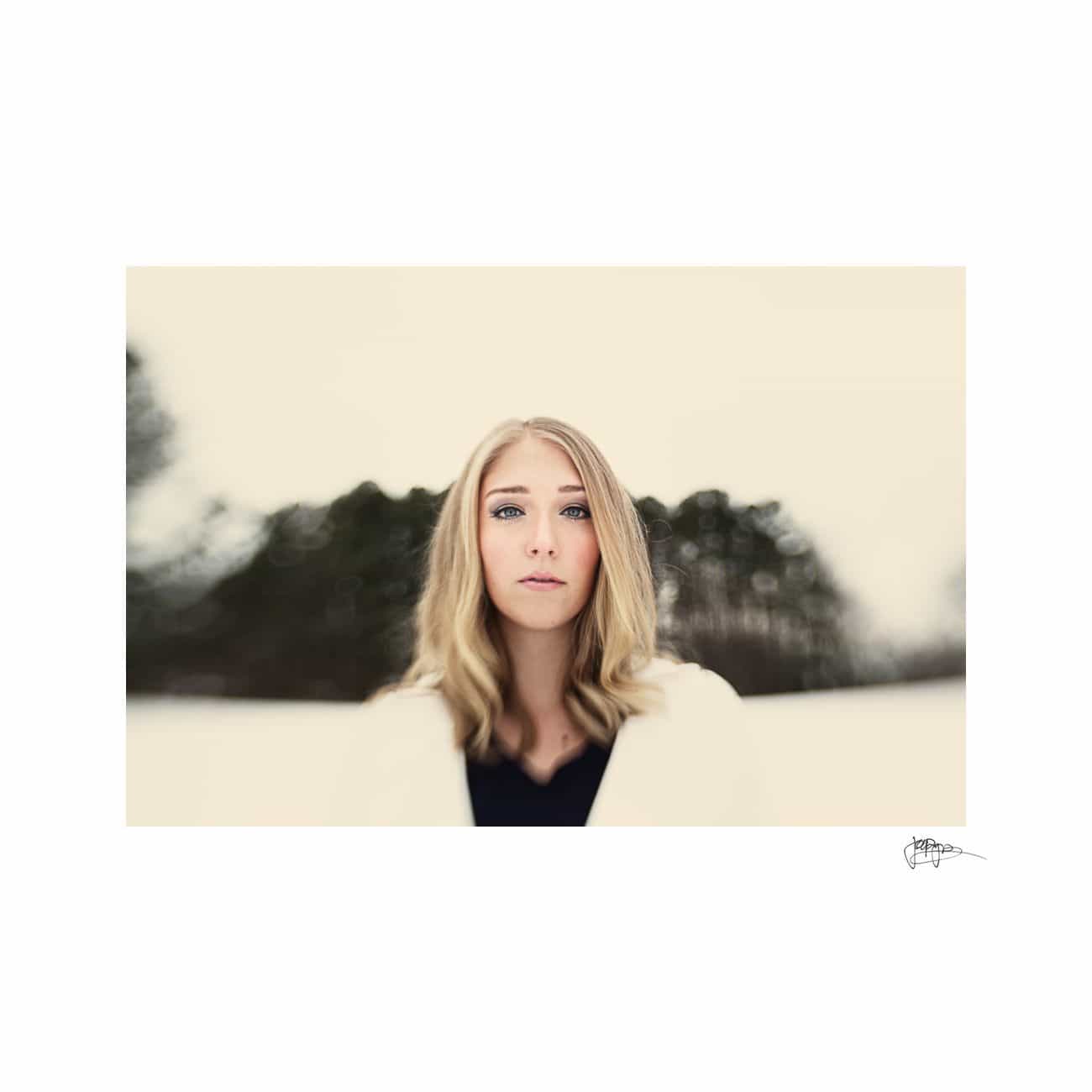 A woman with blonde hair stands outdoors against a blurred background of trees and snow. She is wearing a light-colored coat and dark top, looking directly at the camera with a neutral expression. The image has a soft and serene atmosphere.