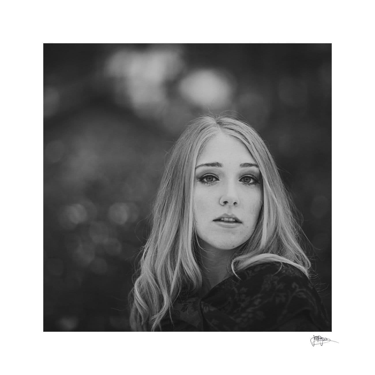 Black and white portrait of a woman with long, light hair, looking directly at the camera. The background is softly blurred, creating a bokeh effect, highlighting the subjects expressions and features.