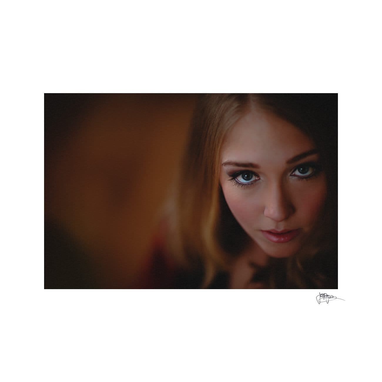 A close-up image of a person with long hair looking directly at the camera. The background is softly blurred, creating a bokeh effect, and the lighting highlights the subjects eyes and facial features.