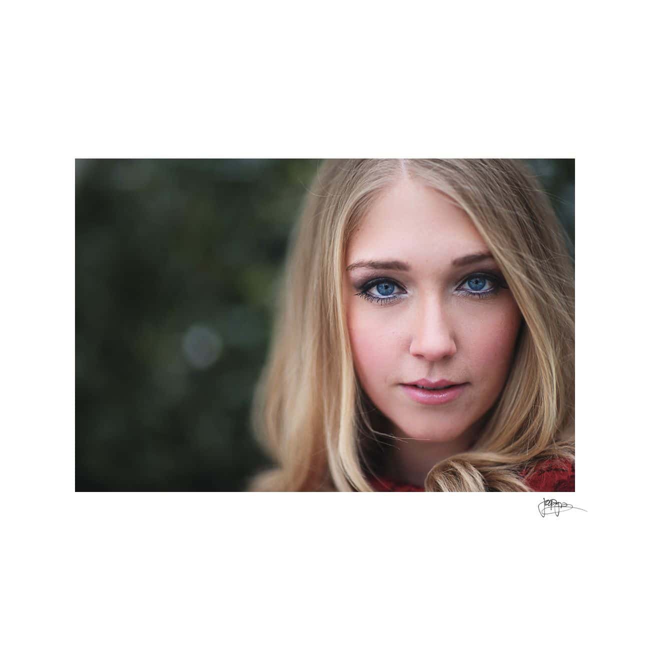 A woman with long blonde hair and blue eyes is looking directly at the camera. She stands against a blurred green background, and her expression is calm and serene.