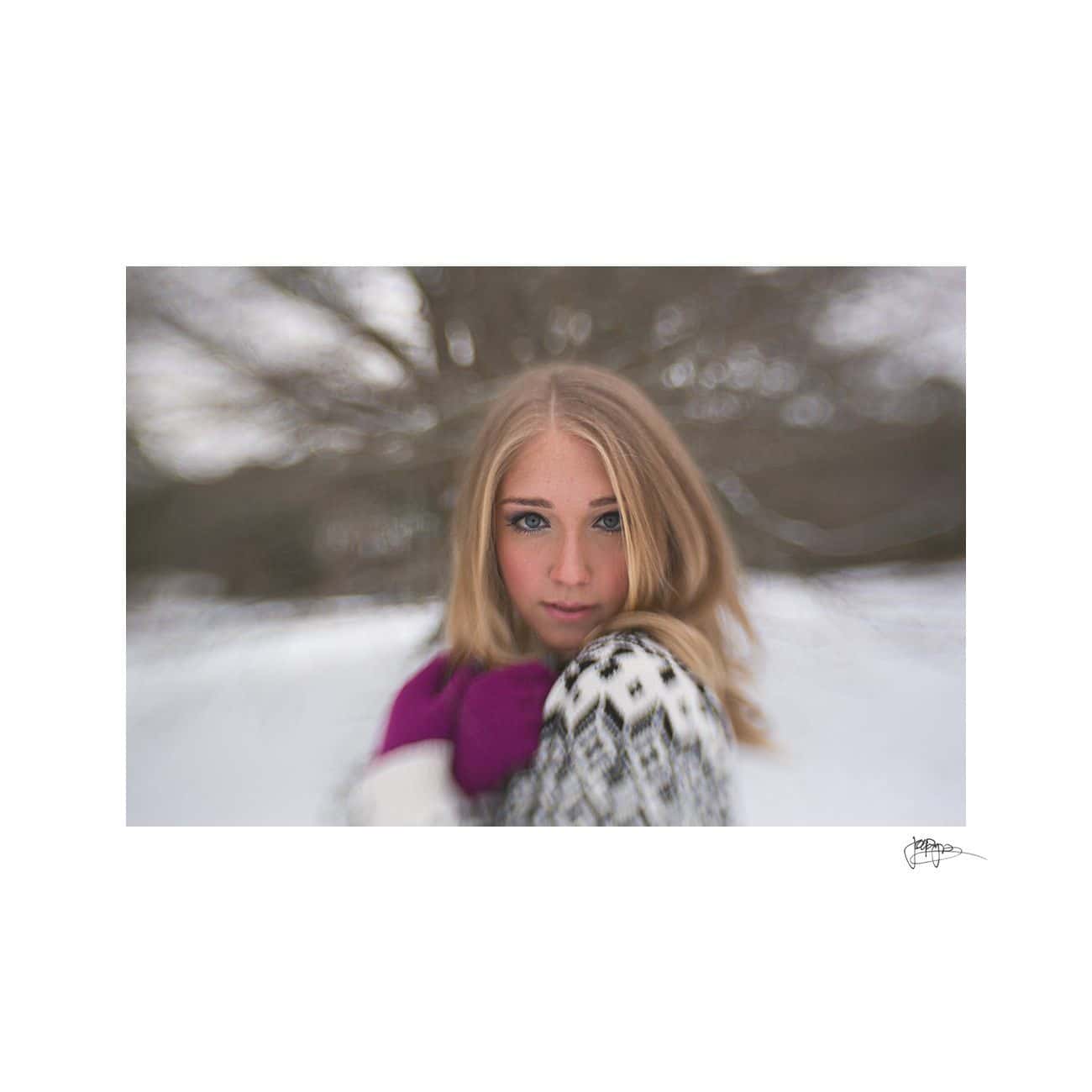 A woman with blonde hair stands outdoors in a snowy setting. She is wearing a patterned sweater and purple gloves, looking at the camera with a blurred background of bare trees.