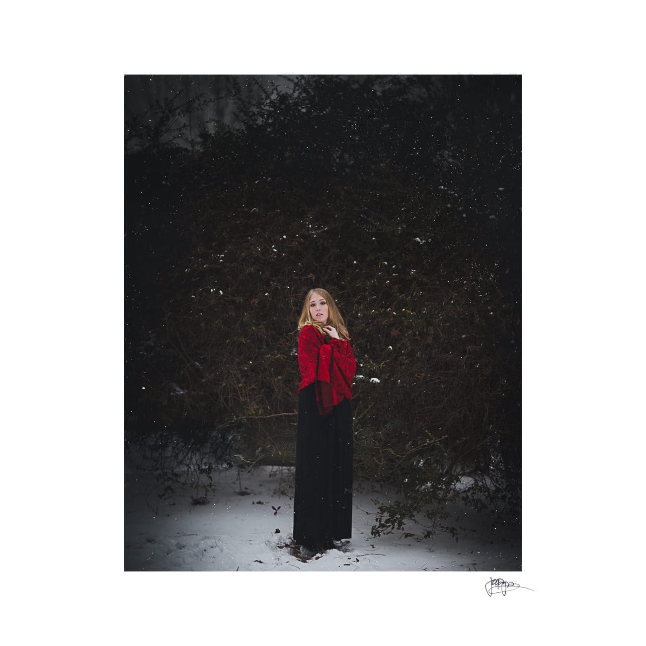 A woman in a red jacket stands in a snowy forest clearing. She is gazing upward, with a dark bushy background behind her. Small snowflakes are falling around. The image has a moody, atmospheric feel.