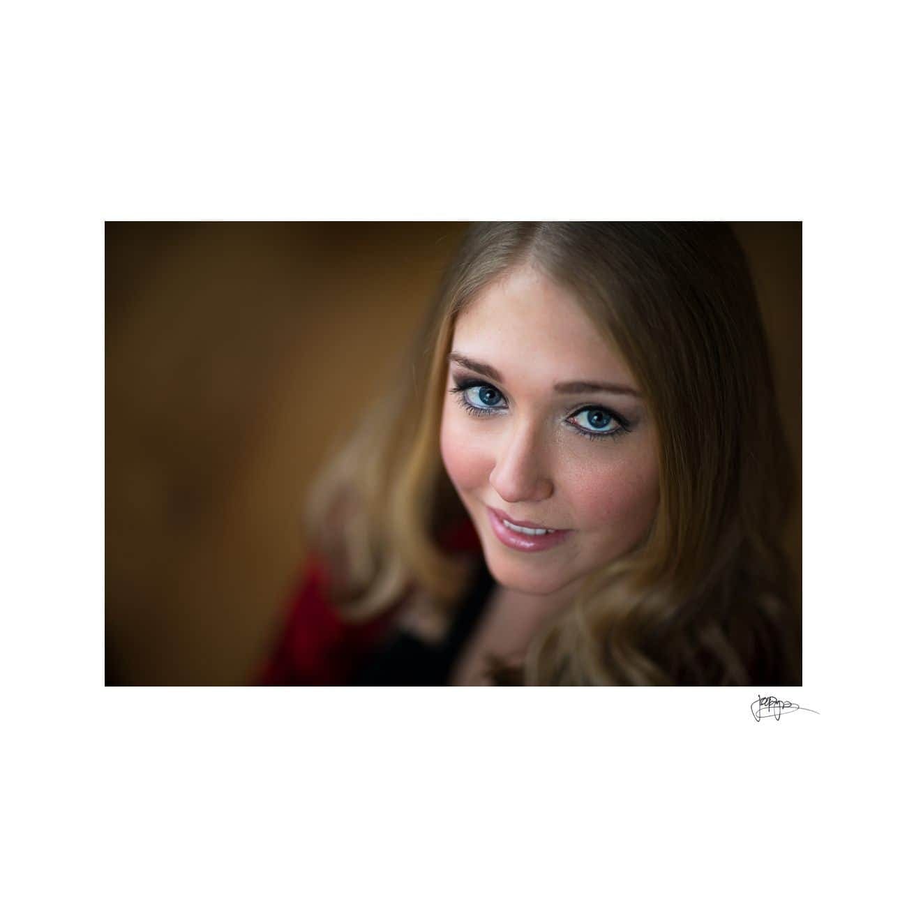 Close-up portrait of a person with long light brown hair and blue eyes, smiling softly and looking up at the camera. The background is softly blurred.