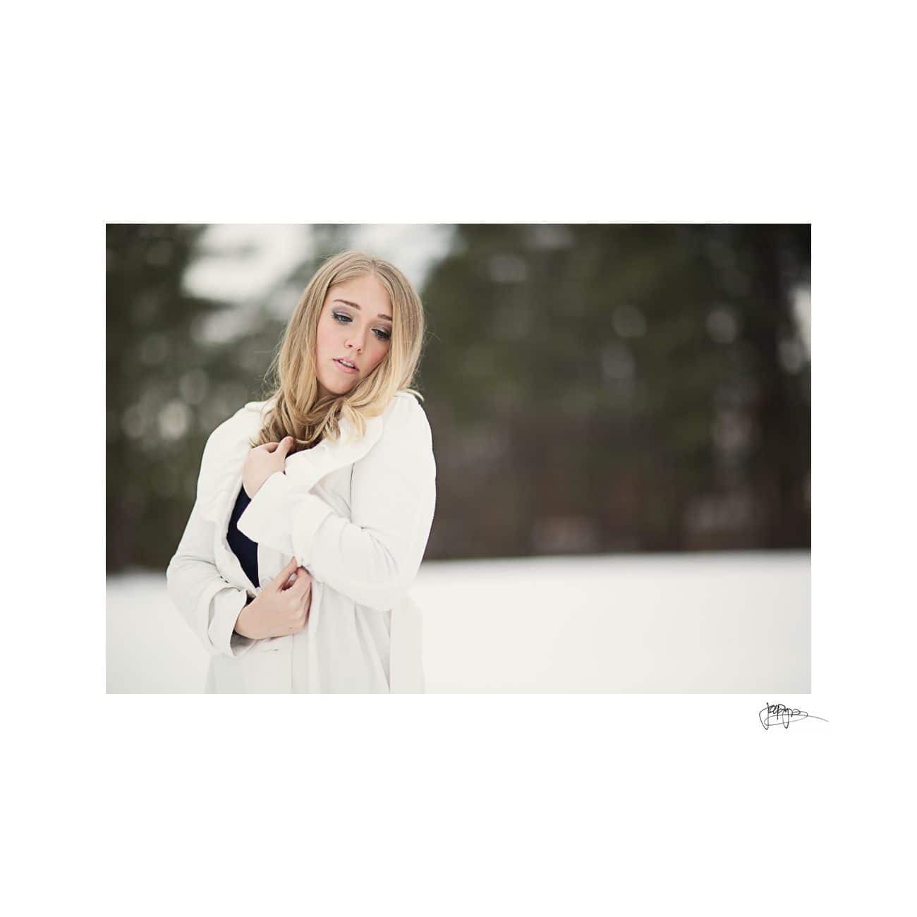 A woman stands outdoors in a snowy landscape, wearing a white coat. The background is blurred with dark green trees, giving a serene winter feel.