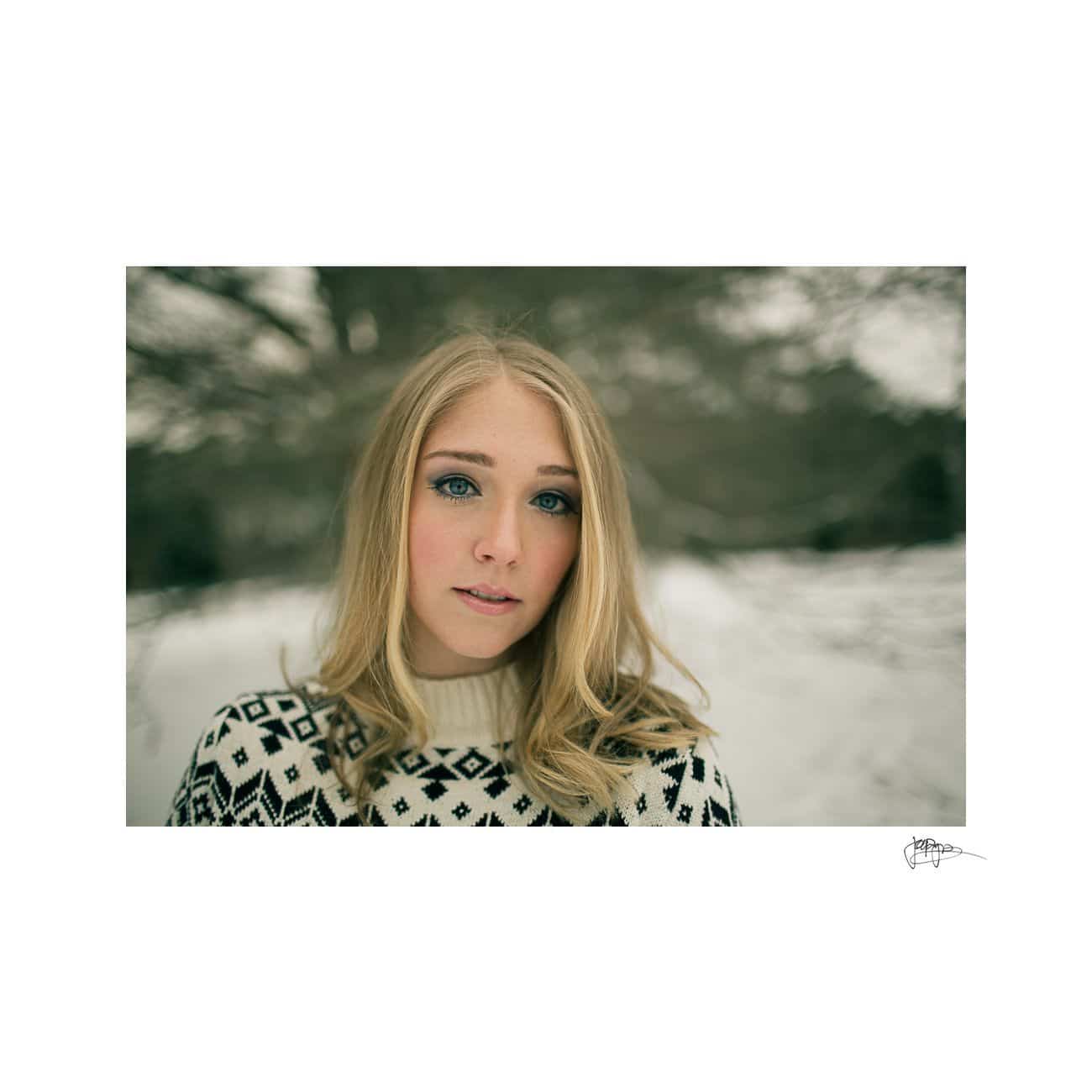 A woman with long blonde hair and blue eyes stands outside in a snowy landscape. She wears a white sweater with black geometric patterns, and the background features blurred trees covered in snow.