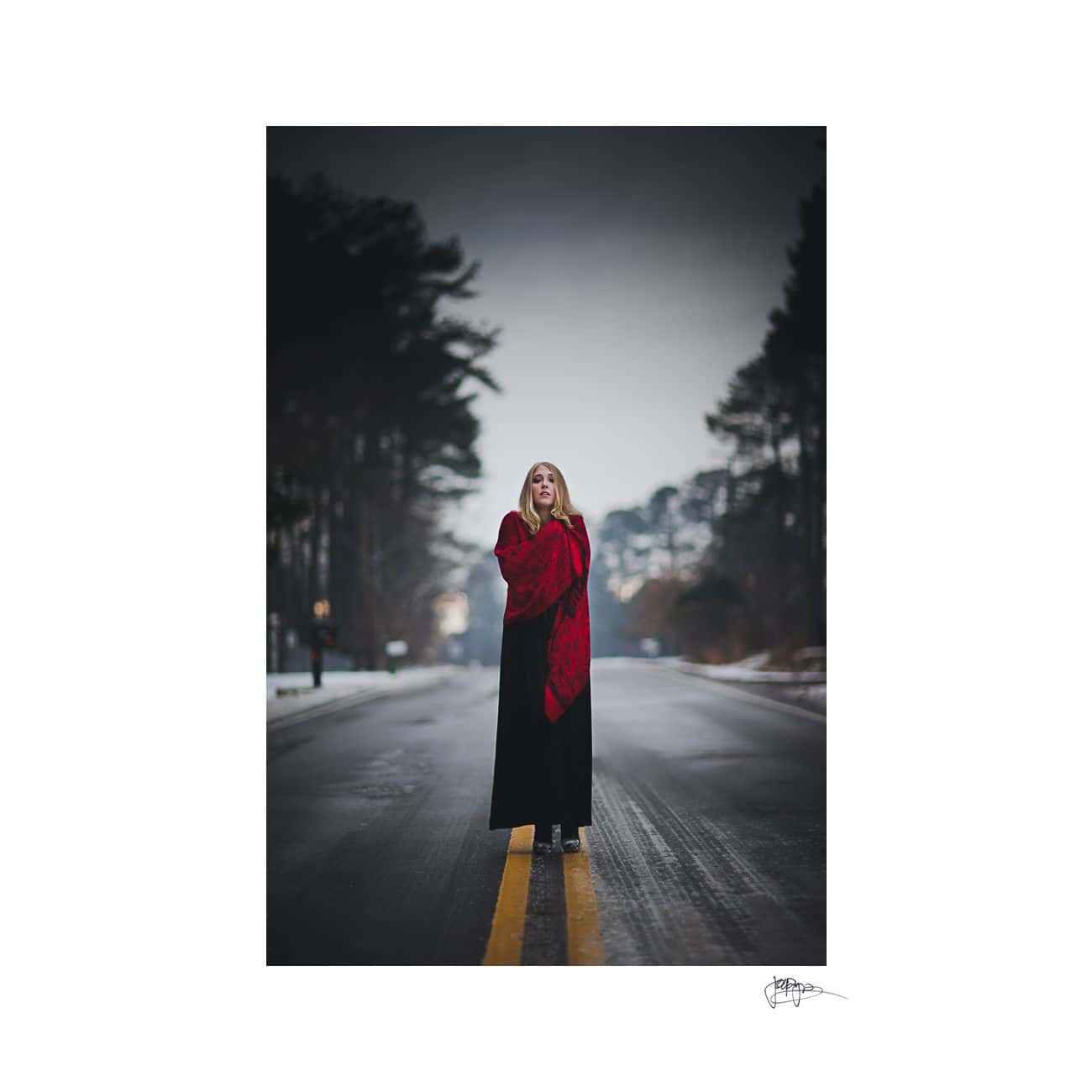A woman stands on a road with a red shawl wrapped around her shoulders. The background is a wintry landscape with trees lining the road. The scene is calm and atmospheric, with a soft, overcast sky.