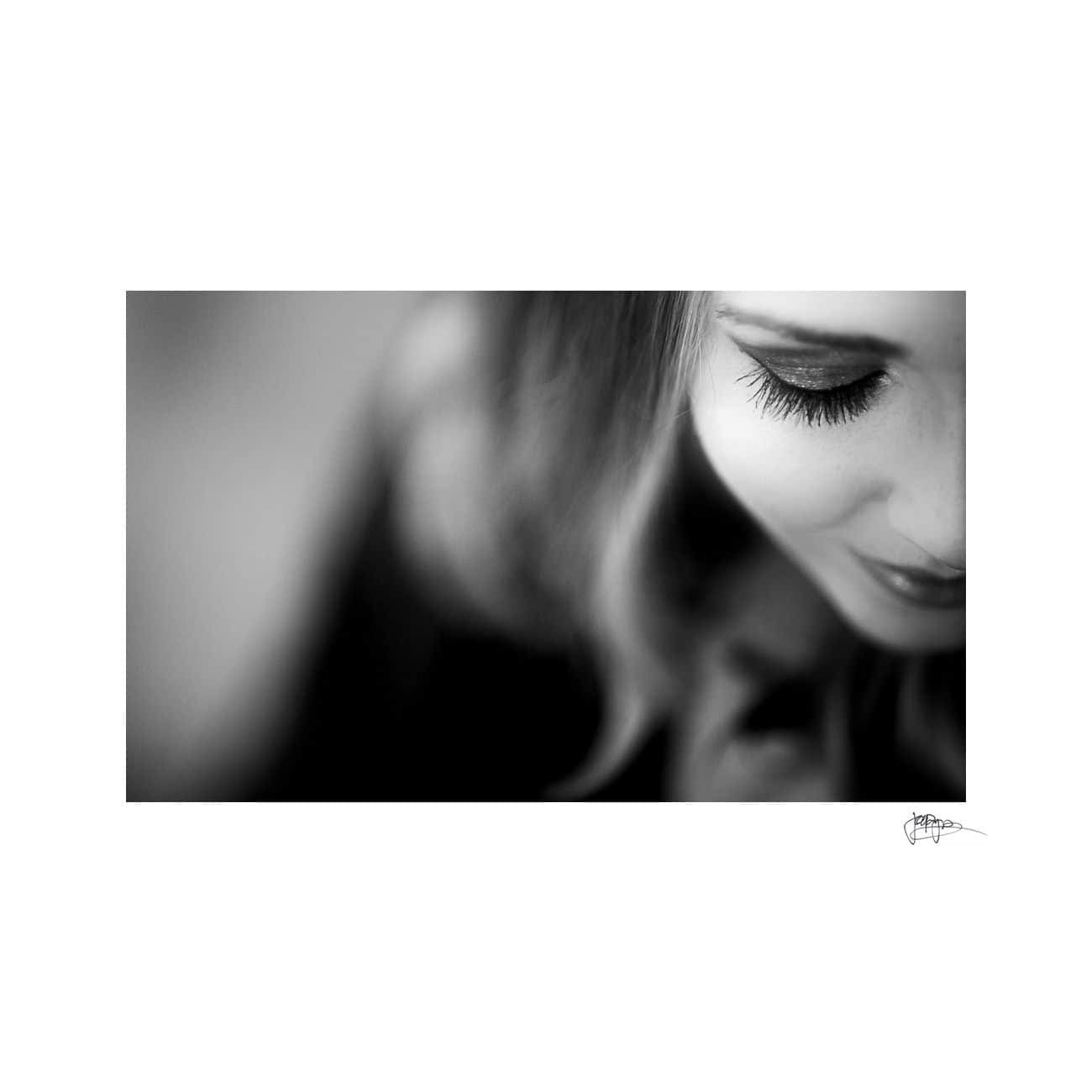 Black and white close-up of a woman looking down, showcasing her eyelashes and a hint of a smile. The background is blurred, focusing attention on her serene expression.