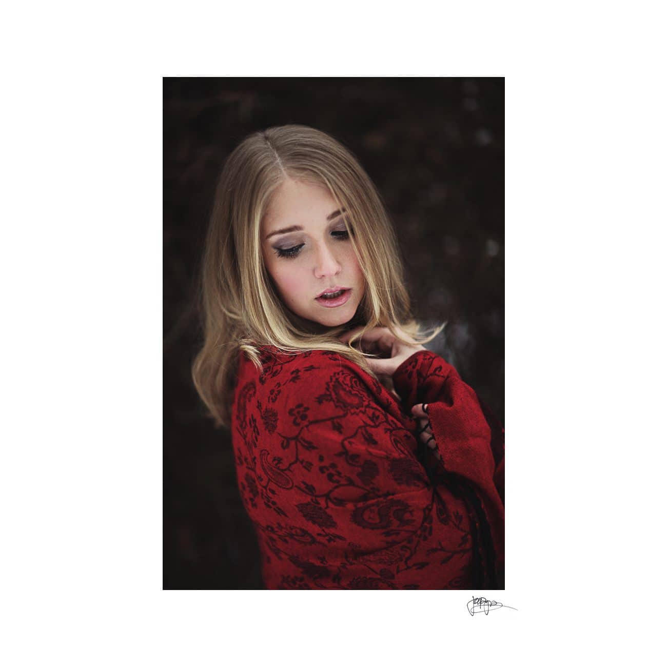 A woman with long blonde hair is wearing a red, patterned shawl. She has her eyes closed and is slightly tilting her head, set against a dark, blurred background.