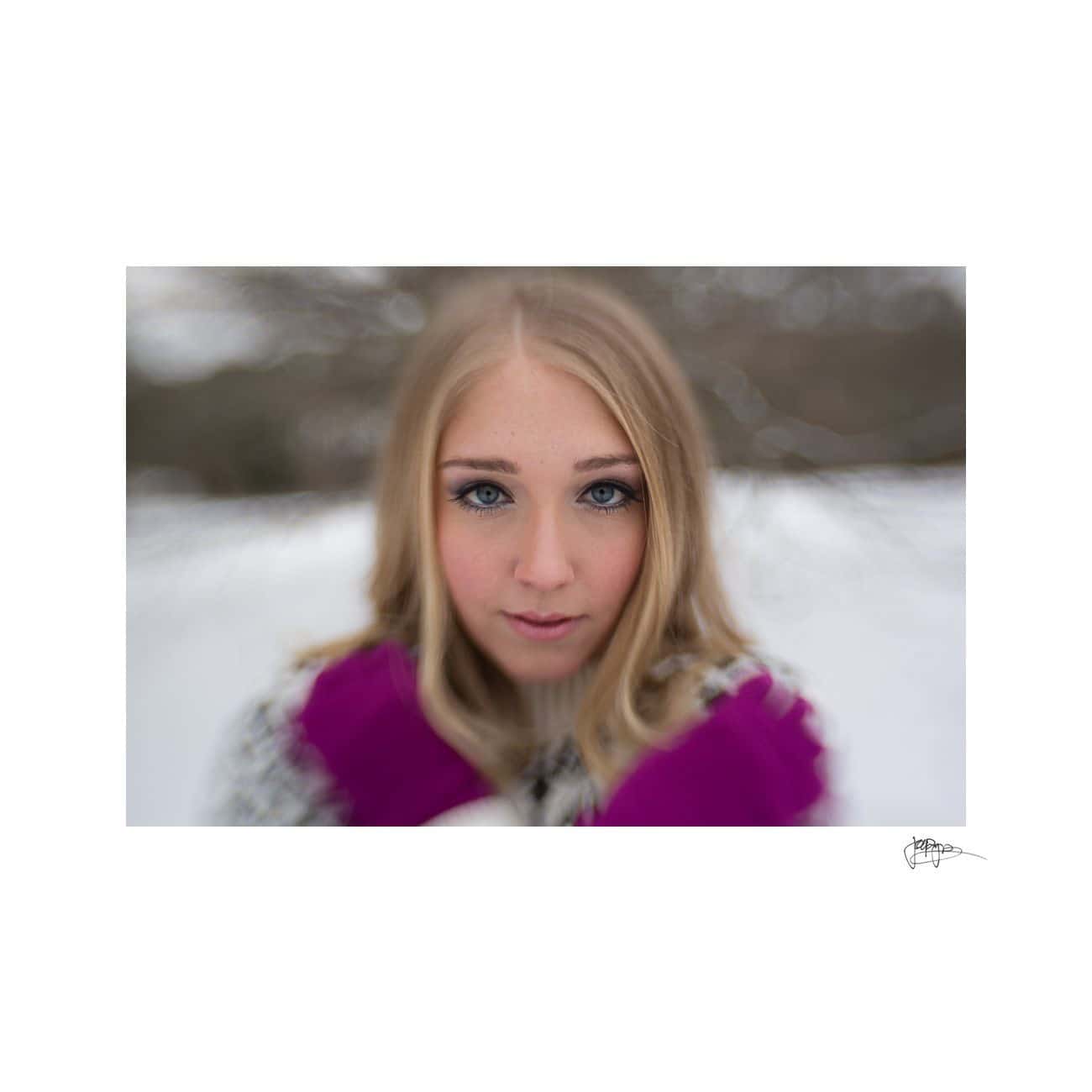 A person with long blonde hair and blue eyes stands outdoors in a snowy setting. They are wearing a black and white coat and pink gloves, with a soft-focus background of trees.