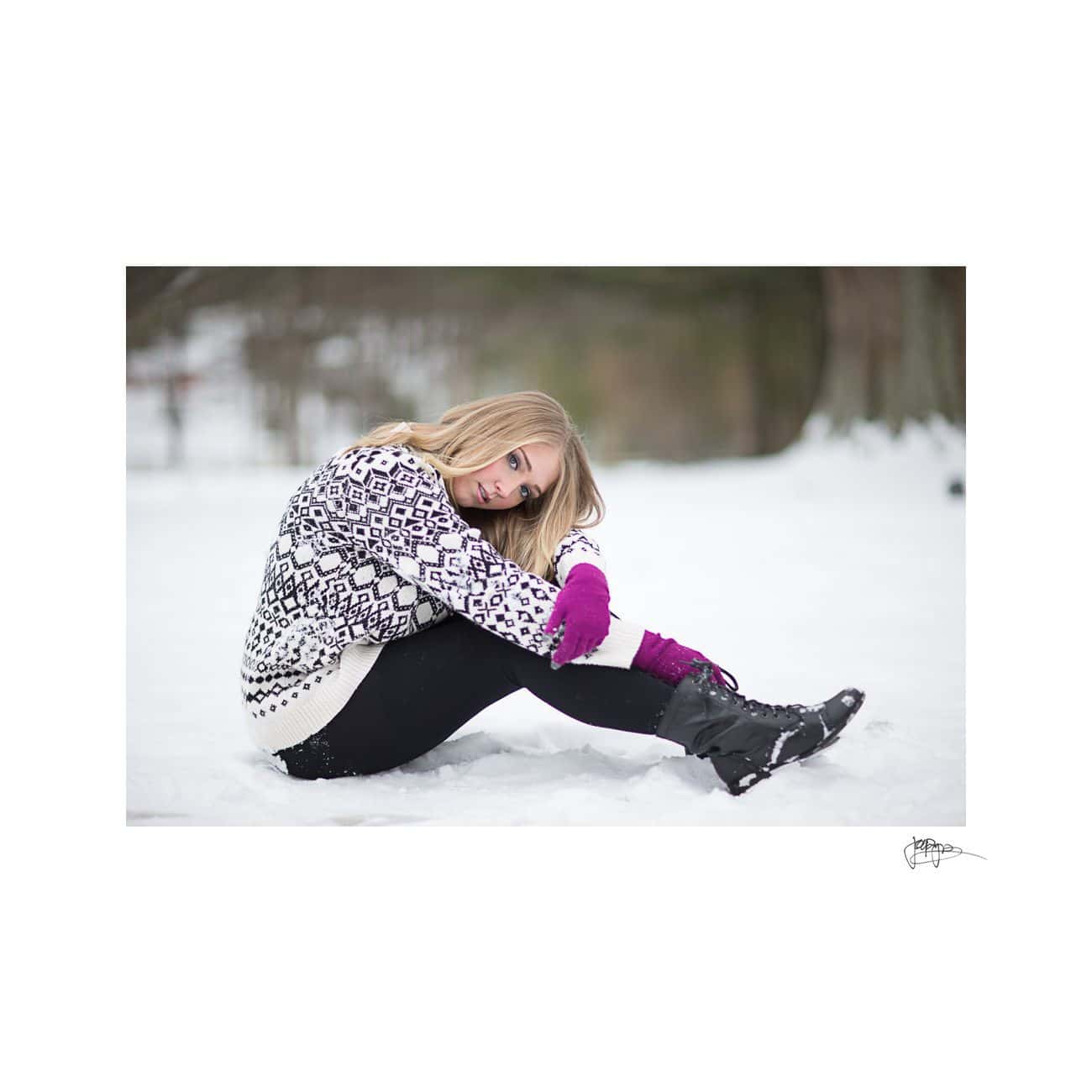 A person sitting in the snow, wearing a black and white patterned sweater, black pants, purple gloves, and black boots. The background is snowy with blurred trees. The person looks relaxed and contemplative.
