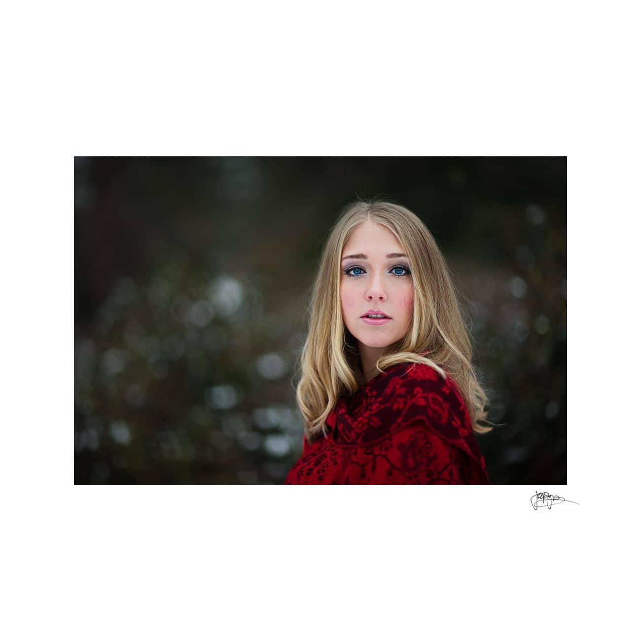 Young woman with long blonde hair stands outdoors, wearing a rich red patterned wrap. The background is a soft-focus mix of green and white, suggesting a natural setting, possibly snowy. Her expression is contemplative.