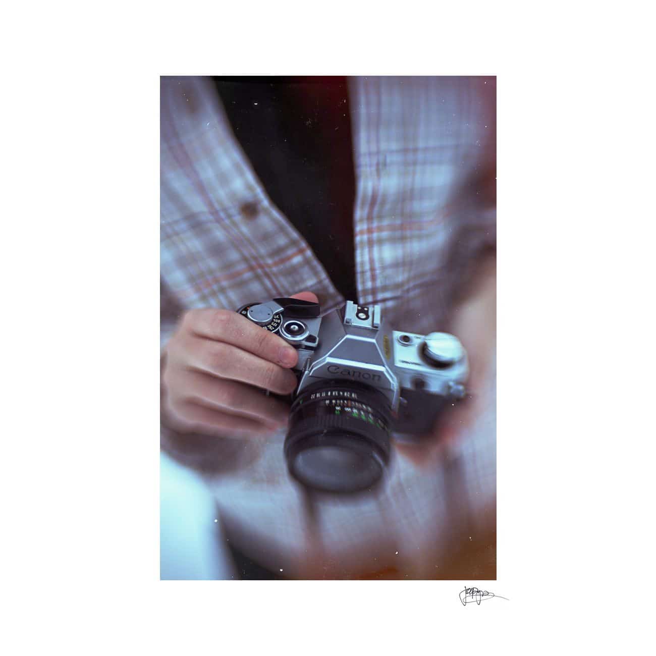 A person in a plaid jacket holds a vintage Canon camera, adjusting the settings with their right hand. The image is slightly blurred, focusing on the camera, giving it a nostalgic feel.