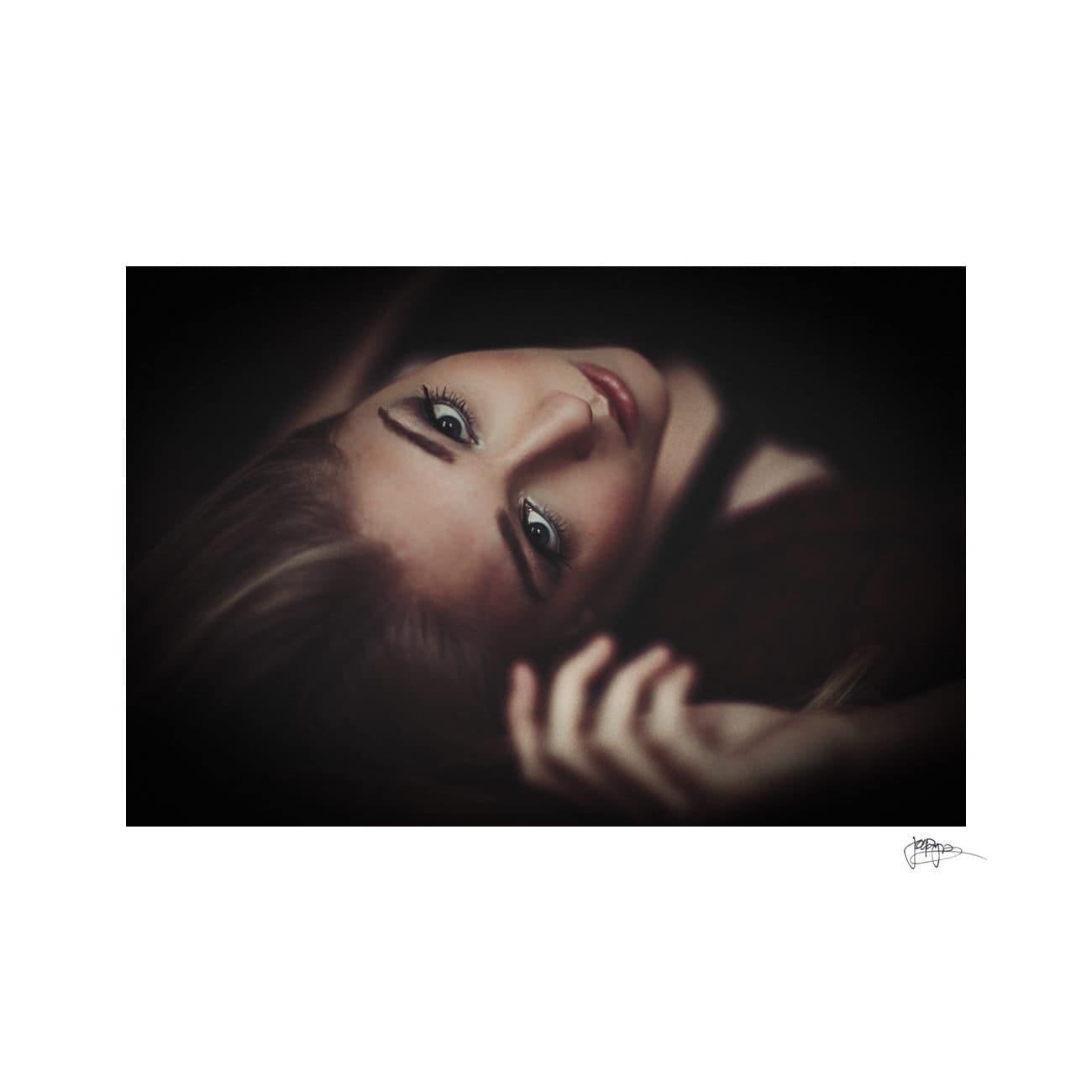 Artistic portrait of a person with long hair lying down, head tilted back. The focus is on their eyes and face, set against a dark background, creating a dramatic and mysterious atmosphere.