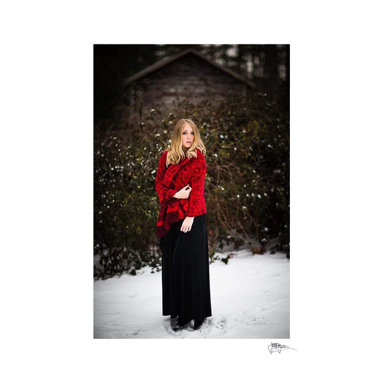 A woman stands in a snow-covered landscape, wearing a long black dress and a red shawl. She is surrounded by greenery and an old wooden shed is in the background. The atmosphere is serene and wintery.